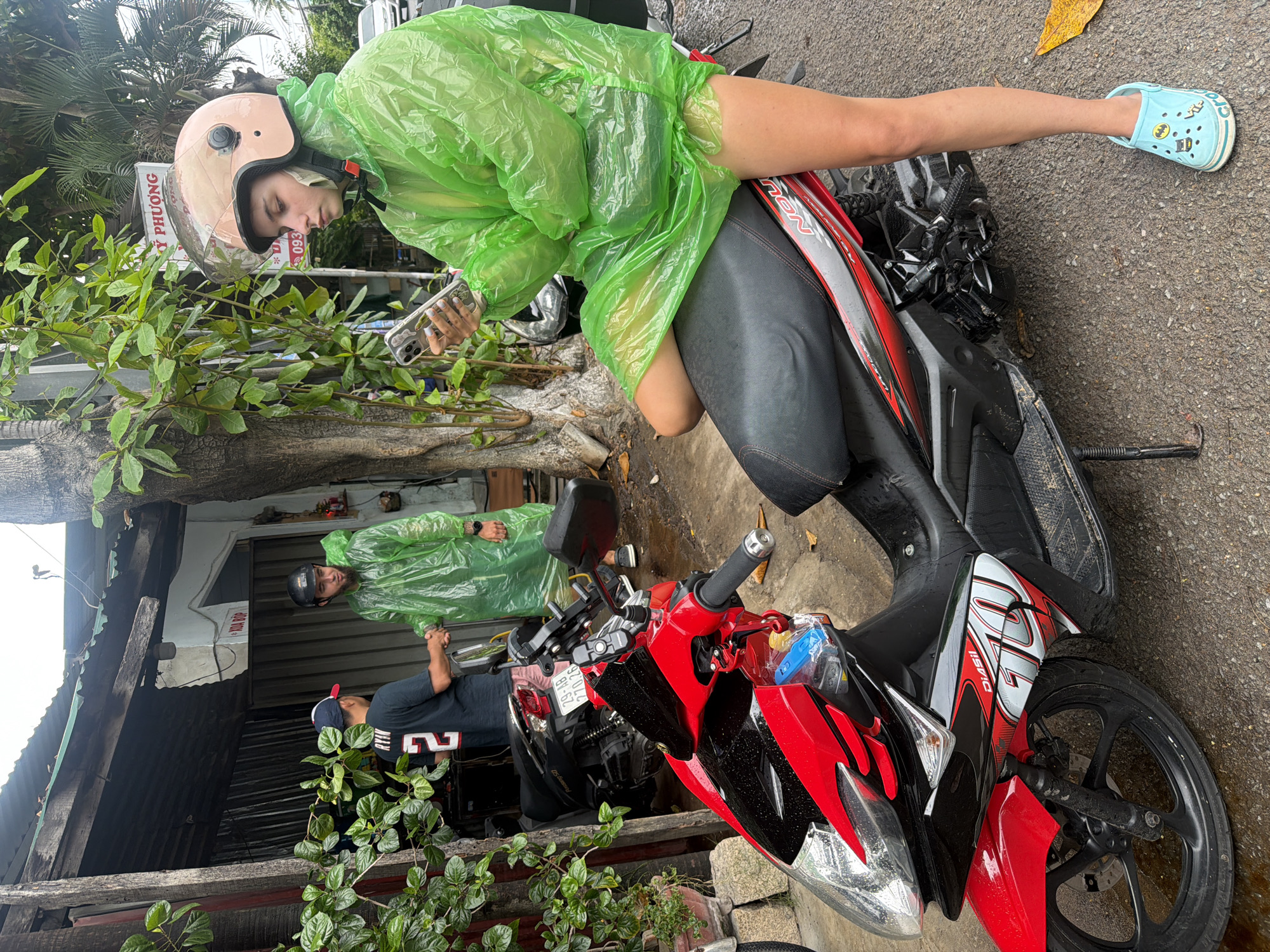 In Nha Trang, Vietnam, on a wet, overcast day, a young woman wearing a light pink helmet and a bright green disposable rain poncho sits on the seat of a red and black motorcycle. She is holding a smartphone in her left hand and appears to be checking it. Her right leg is extended forward, and she is wearing light blue Crocs with Batman decals. Behind her, two men are standing near a rustic structure with a corrugated metal roof. One man, wearing a black shirt with the number "2" on it and a baseball cap, is shaking hands with another man who is wearing a green disposable rain poncho and a black helmet. The ground is wet and appears to be asphalt. A red motorcycle is parked in the foreground. There is foliage and trees surrounding the area. A sign with Vietnamese writing is visible in the background.