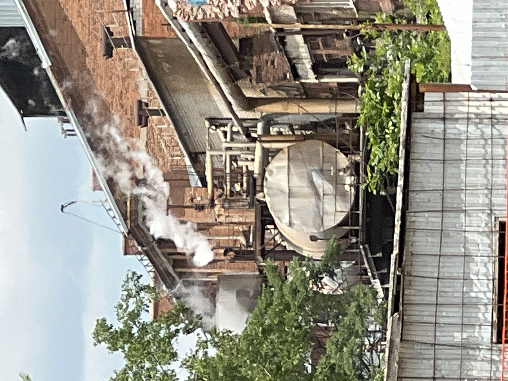 A close-up shot captures an industrial complex under a cloudy sky. Prominently featured are brick buildings with multiple levels and exposed pipes. Steam or smoke billows from several points, indicating active industrial processes. Large cylindrical tanks and various metal structures are visible, suggesting manufacturing or processing facilities. Green foliage encroaches on the lower sections of the buildings and in the foreground, contrasting with the industrial elements. A person in a light-colored suit is partially visible among the trees, possibly a worker or observer. The overall scene depicts a working industrial site with signs of operation and the presence of nature.