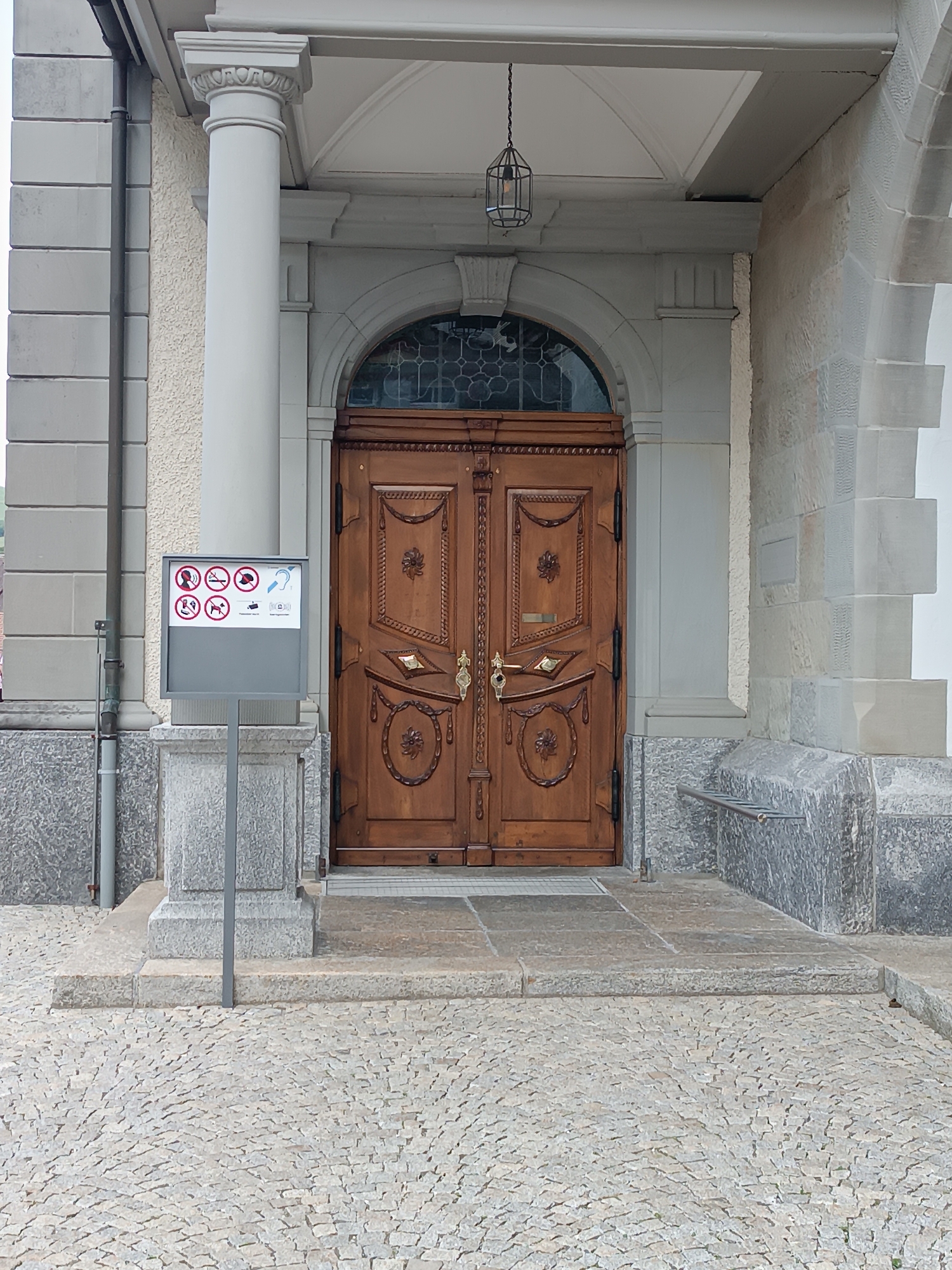 The image shows the entrance to a building in Appenzell, Switzerland. The entrance features a large, ornate wooden double door with intricate carvings. Above the door, a decorative glass window is visible. To the left of the door, a sign with several pictograms, including those prohibiting loud noises and dogs, is displayed on a gray stand. The entrance is flanked by a large white column on the left and a stone facade on the right. The ground in front of the entrance is paved with cobblestones, and there are stone steps leading up to the door. A hanging lantern is suspended from the ceiling above the entrance. The overall impression is one of a historic and well-maintained building.