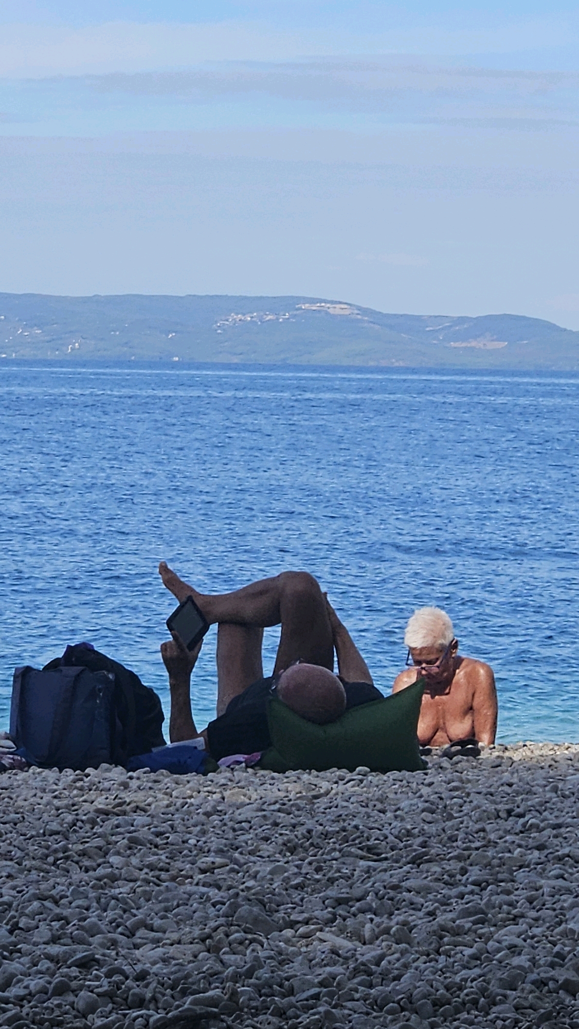 Two people are relaxing on a pebble beach in Općina Podgora, Croatia. The water is a vibrant blue, with a distant mountain range on the horizon. In the foreground, a person is lying on their back, propped up by a green pillow. Their legs are extended, and they are holding a tablet in their right hand, with their thumb positioned as if operating it. A dark blue bag is to their left. To the right of the pillow, a person with short, white hair and glasses is sitting up, facing the water, with their upper body exposed. They appear to be looking down. The beach is covered in grey pebbles.