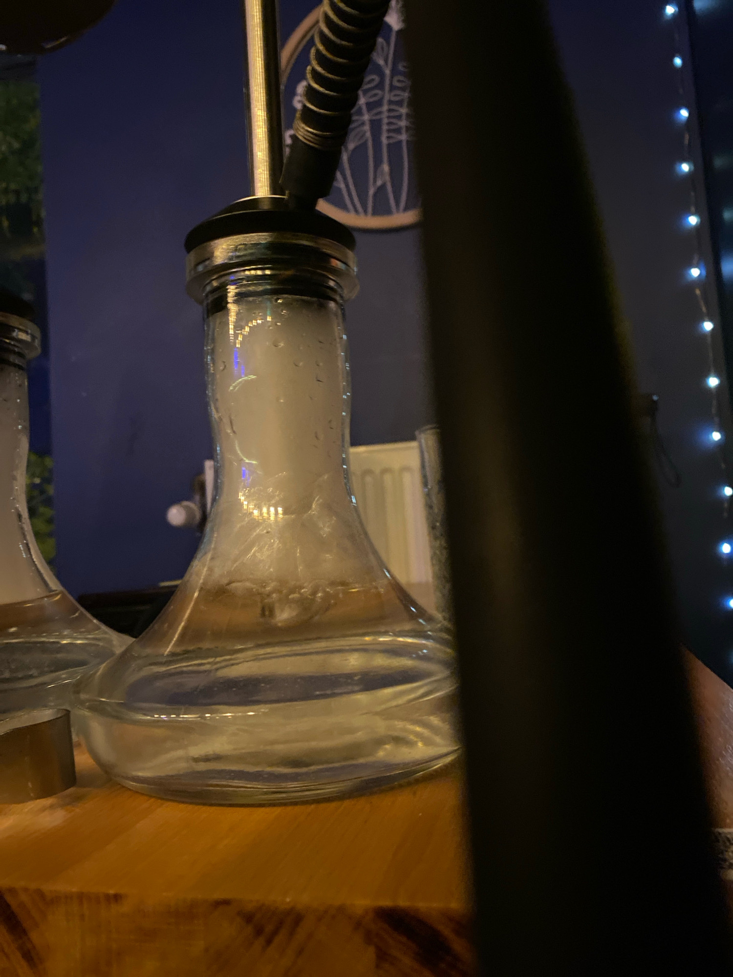 The image shows a close-up of a hookah with smoke rising from its base, suggesting recent use. The hookah is placed on a wooden surface, possibly a table or bar top, in what appears to be an indoor setting. In the background, a blue wall is visible, decorated with a circular motif. String lights with blue LEDs are draped along the right side of the frame, adding to the ambiance. The scene is dimly lit, with the smoke and the reflective surfaces of the hookah catching the light. The overall impression is one of a relaxed, possibly social, environment. There is no visible indication of the specific location within Gliwice, Poland.