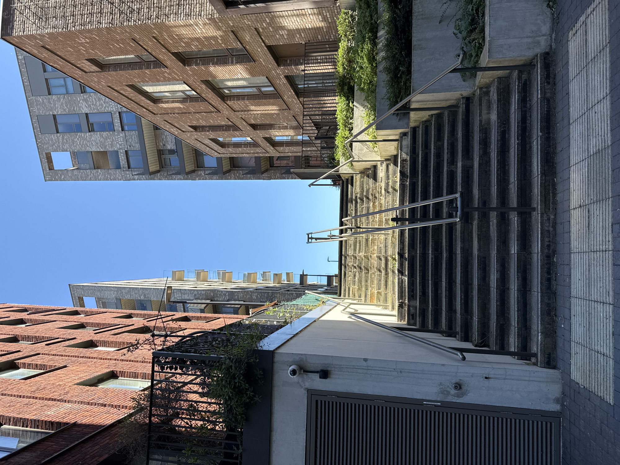 The image captures an upward view of a modern urban passage or alleyway, featuring a steep concrete staircase ascending between multi-story buildings under a clear, bright blue sky. The scene is well-lit, indicating sunny weather during the mid-morning or early afternoon.
In the foreground, a wide flight of dark grey concrete steps leads upwards, flanked by sleek metal handrails on both sides. To the immediate right of the stairs, a concrete wall supports a dense bed of green foliage, possibly a planter. On the left side, another concrete wall with a black metal grille or fence is also adorned with greenery. A small, white security camera is mounted on this concrete wall, with an unreadable white label positioned beneath it. The ground at the foot of the stairs is paved with distinctive textured or patterned tiles.
The passage is defined by contemporary buildings. On the left, a tall structure constructed from reddish-brown bricks exhibits a textured facade with numerous recessed rectangular windows. The building on the right, partially visible behind the foliage and concrete, appears to be of a lighter, greyish-brown brick with prominent windows and angular architectural elements. Further in the background, a third, taller building with a grey and white facade and distinct roof-level structures stands against the sky. The architectural style suggests a modern development, common in urban areas like Greater London, focusing on clean lines and a mix of brick and concrete. No people or specific activities are depicted in the scene.