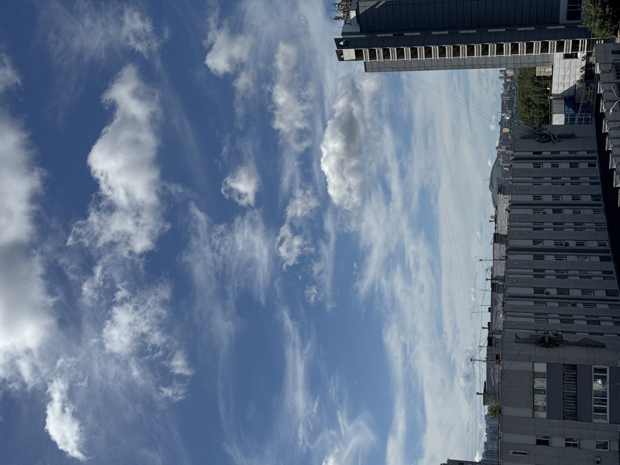 The image presents a view of the city of Krasnoyarsk, Russia, on a partly cloudy day. The sky is a vibrant blue, dotted with various types of clouds, ranging from wispy cirrus formations to more substantial cumulus clouds. In the foreground, several apartment buildings are visible. The building on the right is a tall, grey structure with visible balconies or window frames. To its left, a lower, longer grey building with multiple rows of windows is prominent. Further in the distance, a cityscape with more buildings, some appearing to be under construction, and lush green trees can be seen. A large hill or mountain range forms the backdrop of the distant city. The overall impression is that of an urban environment under a dynamic sky.