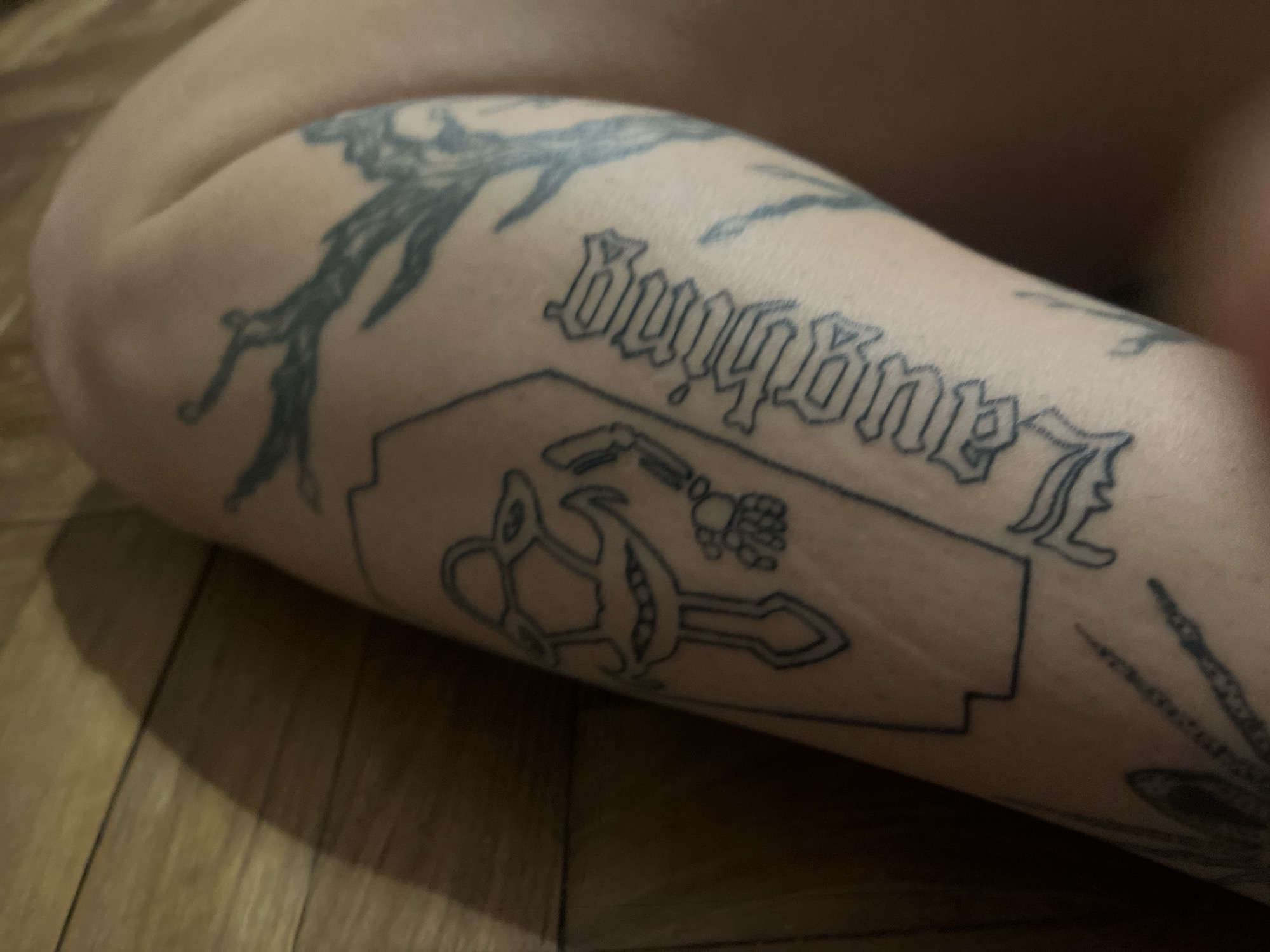 The image provides a close-up view of a person's limb, likely a leg or arm, resting on a wooden floor. The limb is adorned with several distinct tattoos.
The most prominent tattoo is the word "Laughing" written in a decorative, gothic-style script, oriented upside down from the viewer's perspective. Below this text, a larger tattoo forms the outline of a razor blade. Inside this outline, an intricate design is visible, featuring a stylized object that resembles a key or an axe, incorporating details that look like an eye or a pod of peas, and a small, bony hand or a bomb with a fuse.
Other tattoos are partially visible on the limb. Above the "Laughing" text, darker, abstract or organic patterns, possibly depicting tree branches, extend across the skin. Towards the bottom right of the frame, a hint of another tattoo with radiating lines, possibly a sun or starburst design, can be seen.
The background consists of a warm-toned wooden floor with visible planks, suggesting an indoor setting. The lighting is dim, contributing to a subdued atmosphere. The image focuses entirely on the tattooed limb, offering no further details about the person or any ongoing activity other than the display of the tattoos. The setting is Vicente López, Argentina.