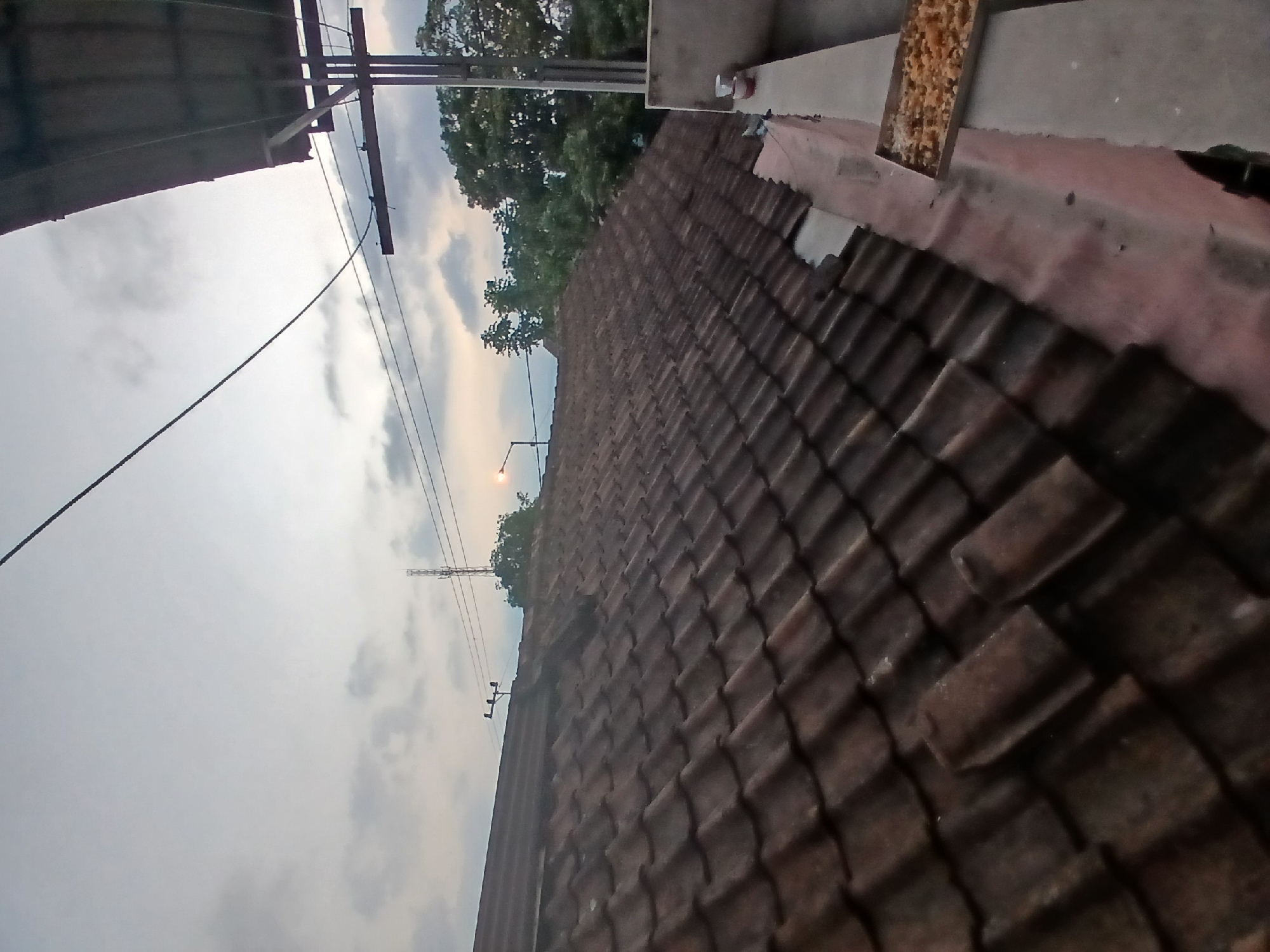 The image shows a view of rooftops and the sky during what appears to be dusk. The foreground is dominated by a weathered, brown tile roof that slopes downwards towards the right. Beyond the roof, the sky is overcast with clouds, tinged with the soft colors of sunset. A telecommunications tower is visible in the distance. Power lines crisscross the sky. To the right, a portion of a structure with a dark, sloped roof is visible, supported by poles. On a concrete ledge in the lower right corner, there is a small tray containing what looks like food, possibly for birds or other animals. The location is identified as Tenggarang, Indonesia.