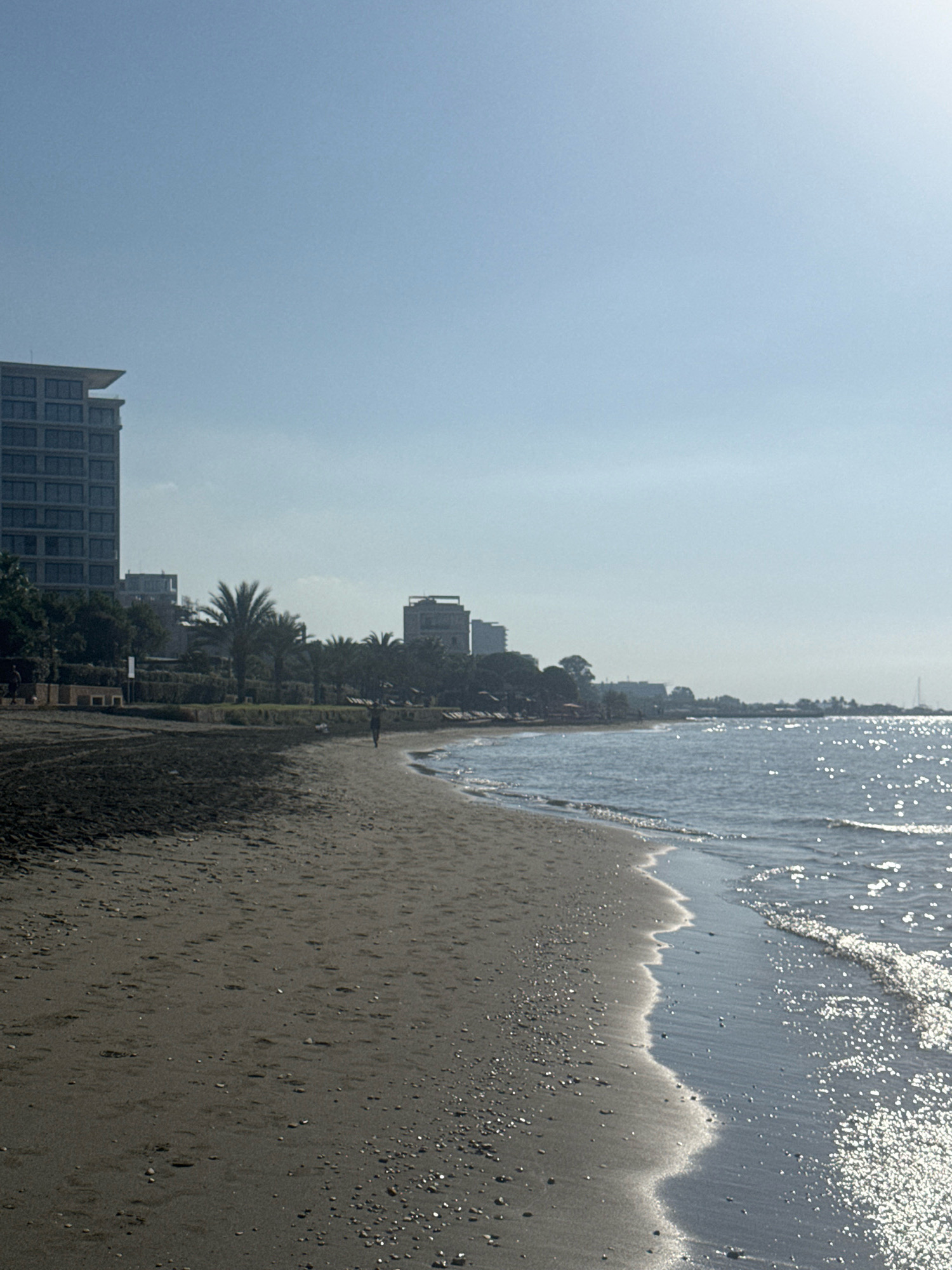 The image shows a sandy beach with the sea on the right and buildings in the background, located in the Agios Tychona Community, Cyprus. The sand is dark and appears wet closer to the water. Small pebbles are scattered across the sand. The sea is calm with gentle waves lapping at the shore, and sunlight reflects off the water's surface, creating a shimmering effect. In the background, several buildings are visible, including a tall, modern structure on the left and other lower buildings further down the coastline. Palm trees are interspersed among the buildings and vegetation. A single person is walking on the wet sand near the water's edge. The sky is clear and pale blue, with a faint haze. The overall scene depicts a serene coastal environment with a clear day.