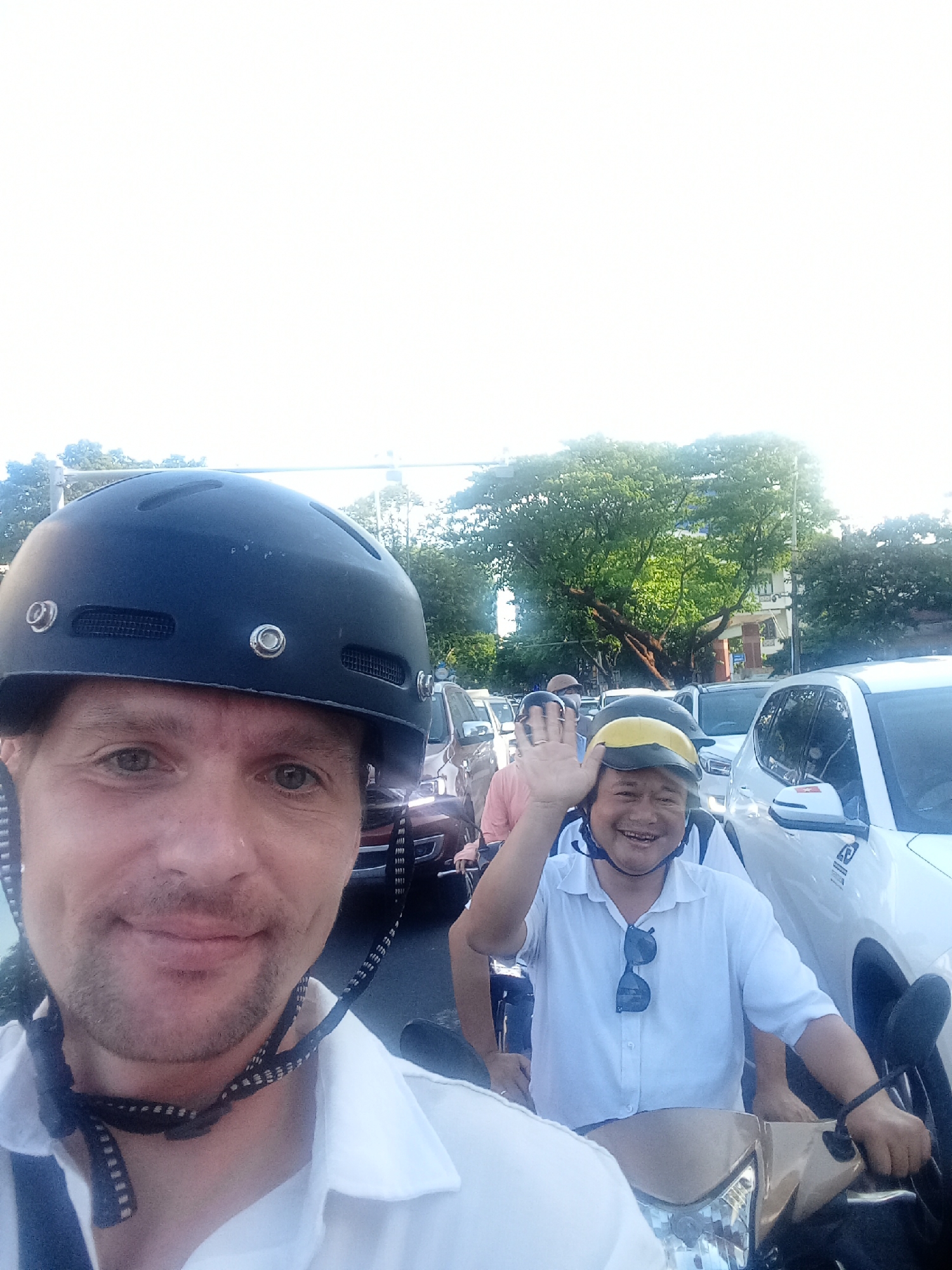 This is a selfie taken on a motorcycle in traffic, likely in Đà Nẵng, Vietnam. Two men are visible. The man in the foreground, on the left, is wearing a black helmet and a white collared shirt. He has light skin, short brown hair, and is looking directly at the camera with a slight smile. The other man, on the right, is also wearing a helmet, this one yellow and black. He is wearing a white collared shirt and smiling broadly, waving at the camera. He has darker skin and appears to be of Asian descent.
They are riding on a motorcycle, as indicated by the handlebars and front of the vehicle visible in the lower part of the image. The background shows a busy street with several cars and other motorcycles, suggesting a traffic jam. Trees and buildings are visible in the distance, with a bright, overcast sky overhead, indicating it is daytime. The overall mood appears to be lighthearted and cheerful, despite being stuck in traffic. There is a Vietnamese flag decal visible on the side mirror of the white car to the right.