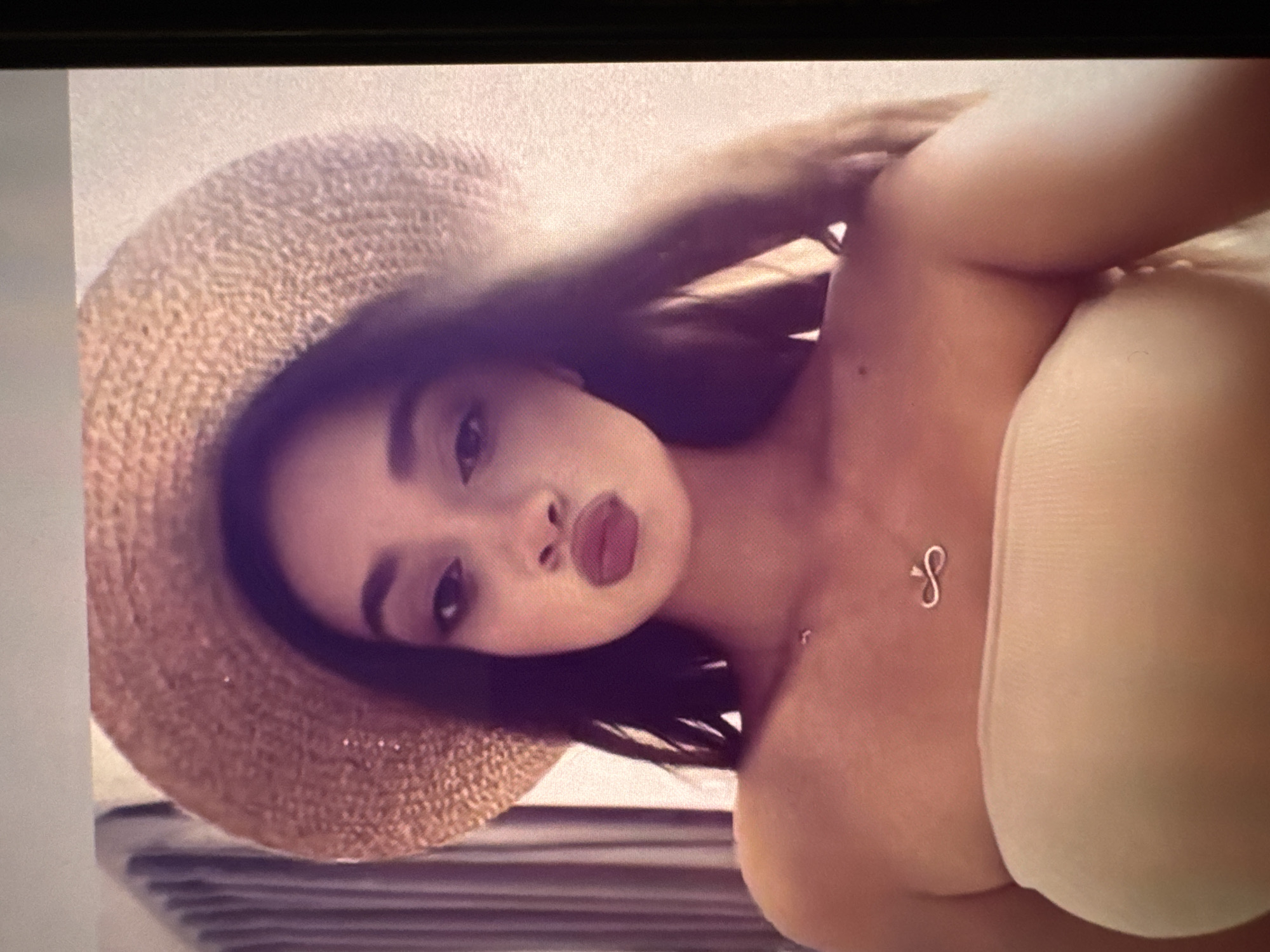 A woman wearing a straw hat and a tube top poses for a selfie. She has brown hair and fair skin, with a silver necklace. The location is not discernible.