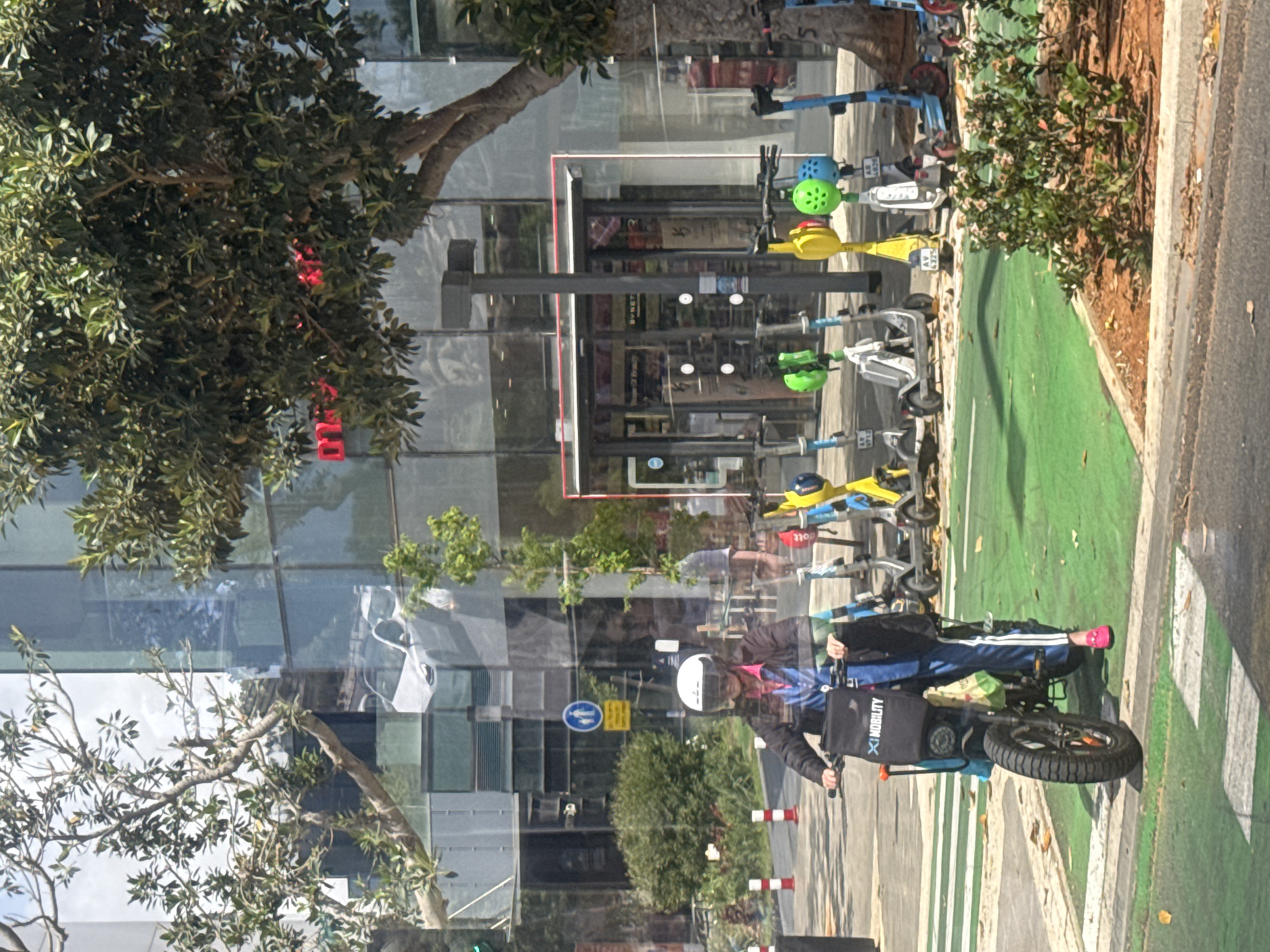 The image shows a street scene in Tel Aviv, Israel, featuring a bicycle lane. A person wearing a helmet and jacket is riding a bicycle with a "Mobility" bag attached to the front. The bicycle is on the green-painted bike lane. Along the lane are rows of electric scooters lined up. Behind the bike lane, there is a modern building with a glass facade. Trees are visible along the sidewalk.