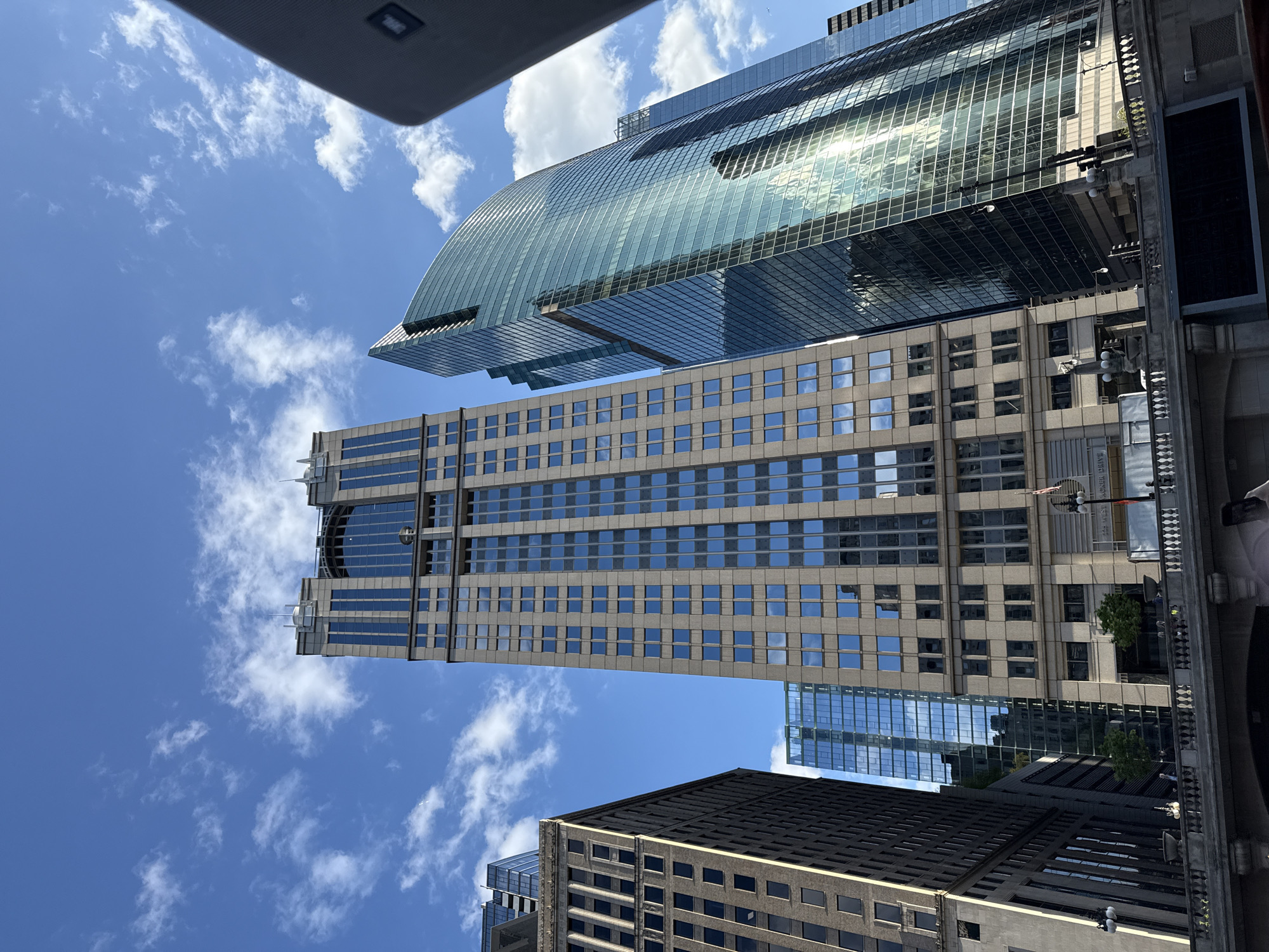 The image shows a view of buildings in Chicago, USA, taken from a low angle. Several skyscrapers dominate the frame, set against a backdrop of a blue sky with scattered white clouds. The central building features a repetitive pattern of windows. A modern glass building with a curved top is partially visible at the top of the frame, reflecting sunlight. Other buildings with darker exteriors are present towards the bottom of the image. A bridge or elevated walkway structure is seen on the right side of the frame, providing context to the street-level perspective. The overall scene captures the urban landscape and architectural diversity of downtown Chicago.
