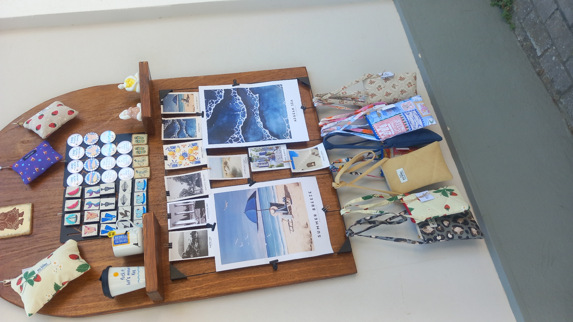 The image shows a wooden display board mounted on a white wall, likely in Kos, Greece, given the presence of tourist-themed items. The board is adorned with various decorative objects and merchandise.
At the top, there are several small, fabric items with strawberry and floral patterns, and one with a blue geometric design. Below these, a collection of round pins or badges is displayed on a black backing. These pins feature nautical motifs like sailboats, fish, and anchors, as well as what appear to be stylized images of a pig. Some pins have text, including "GOOD MORNING". To the right of the pins are two small ceramic figurines, one seated and holding a yellow object, and another that is partially obscured.
On a wooden shelf below the pins, there are two mugs. One is white with a black lid and features the text "fuck it, let's move Kos". The other mug is white and displays a "20% OFF" sticker.
The main part of the display consists of several printed pictures and postcards clipped to the wooden board. These include scenes of beaches, architectural ruins, lemon patterns, and abstract blue wave patterns. Some of these also have Greek text such as "KASTRI KOS", "ASCLEPIEION KOS", "HERAKLEION BRIDGE", "AEGEAN SEA", and "SUMMER BREEZE".
Hanging from hooks below the wooden board are several small bags. These bags have various patterns including leopard print, strawberries, a plain tan color, and a colorful fabric with geometric and illustrated designs.
The wall is white, and the floor visible at the bottom of the image is a gray paved area with some greenery. The overall presentation suggests a retail display of souvenirs or local crafts.