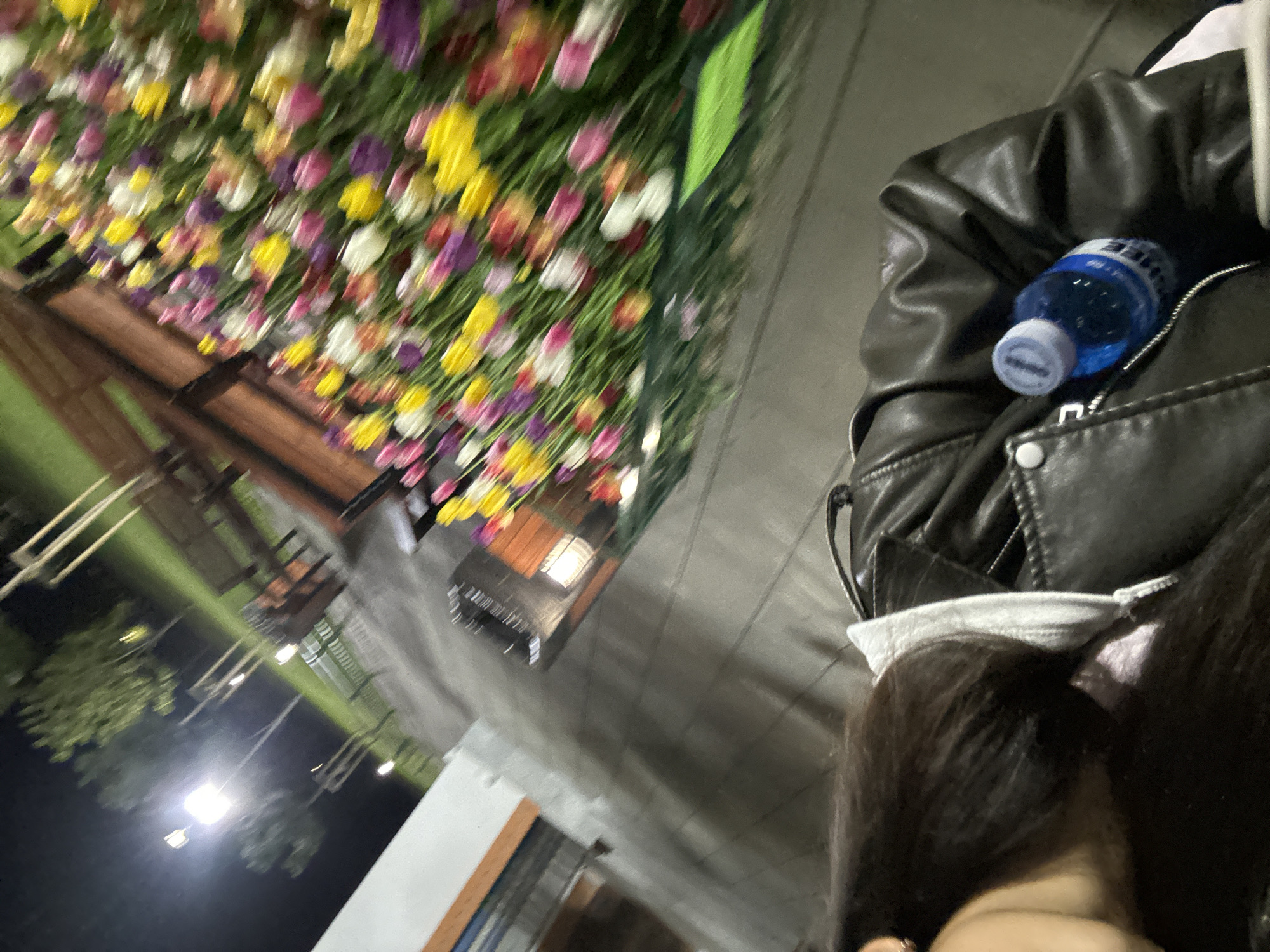 A blurry image shows a person in a black leather jacket with a water bottle tucked into a pocket. The person is wearing a white face mask and has long dark hair. In the background, there's a flower arrangement with purple, yellow, and white flowers. There is an awning structure. The scene appears to be outdoors at night, with lights visible in the background. I cannot determine whether this image is from Sofia, Bulgaria.