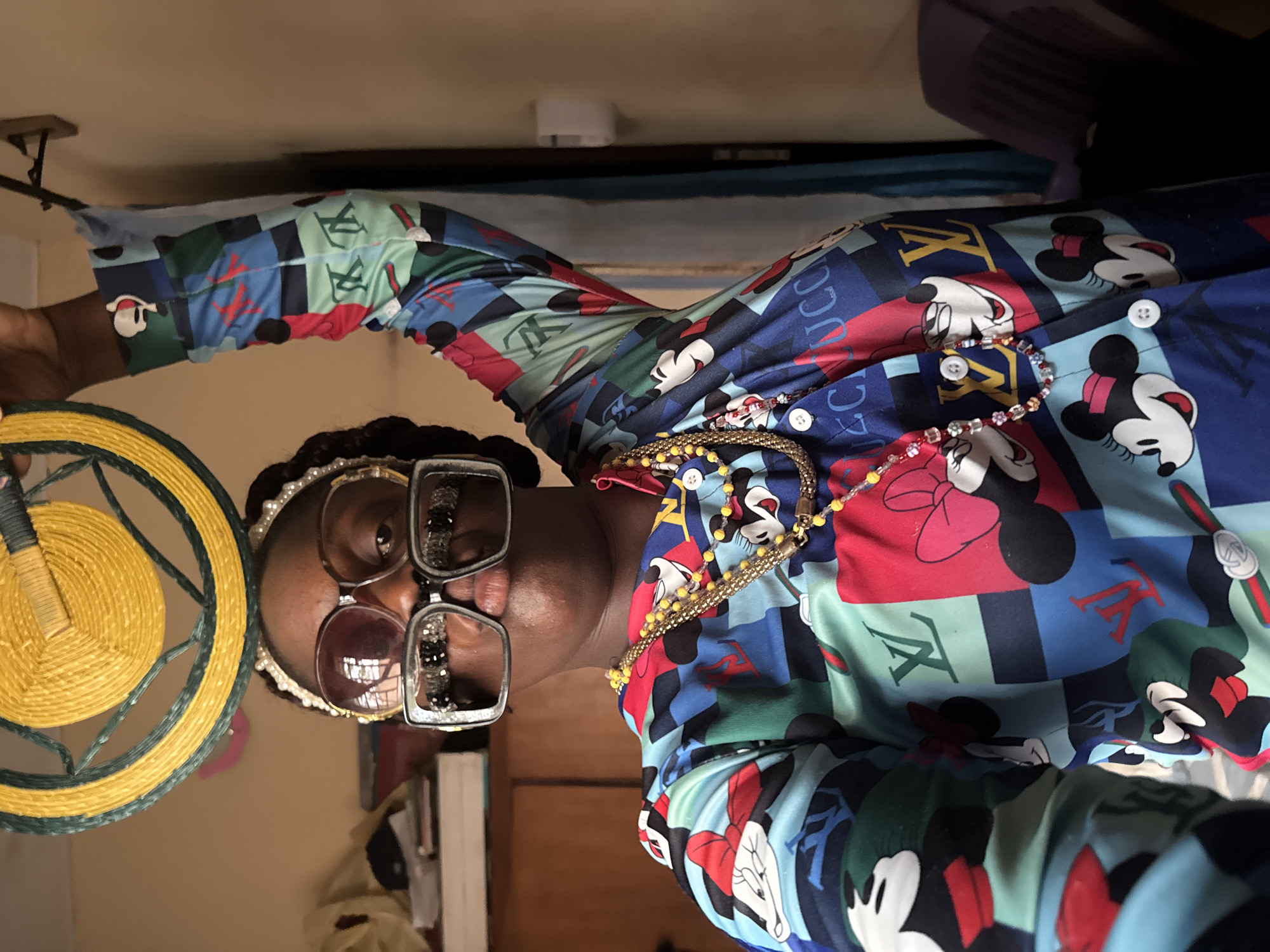 A woman is captured in a medium shot, wearing multiple pairs of glasses, one over the other. She is adorned with multiple beaded necklaces. Her patterned shirt features cartoon figures and the letters "LV" and "Gucci." She holds a round, woven, yellow and blue object in her left hand. The background includes a white bookshelf, a portion of a bed with a blue cover, and beige walls. The location is unspecified, and there is no indication of Madangala, Nigeria.