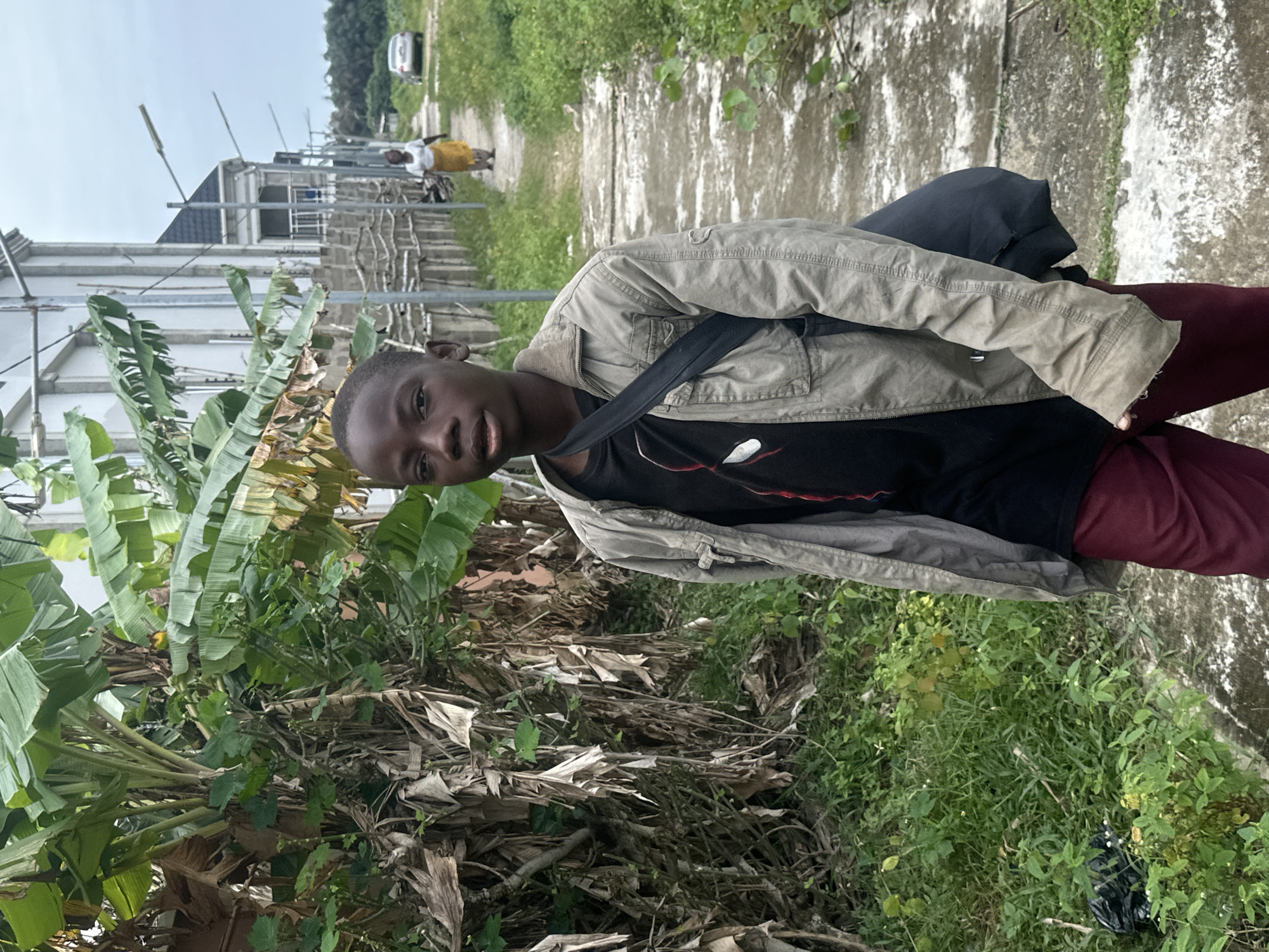 A young boy stands on a concrete pathway in Badagry, Nigeria. He is wearing a light gray jacket over a black t-shirt and maroon pants, with a dark bag slung over his shoulder. The pathway is flanked by lush green vegetation, including banana trees, and leads towards a modern, multi-story building with white walls and a dark roof. In the background, a woman in a white top and yellow skirt walks away from the camera, and a silver car is visible further down the path. The sky is overcast, suggesting a cloudy day.