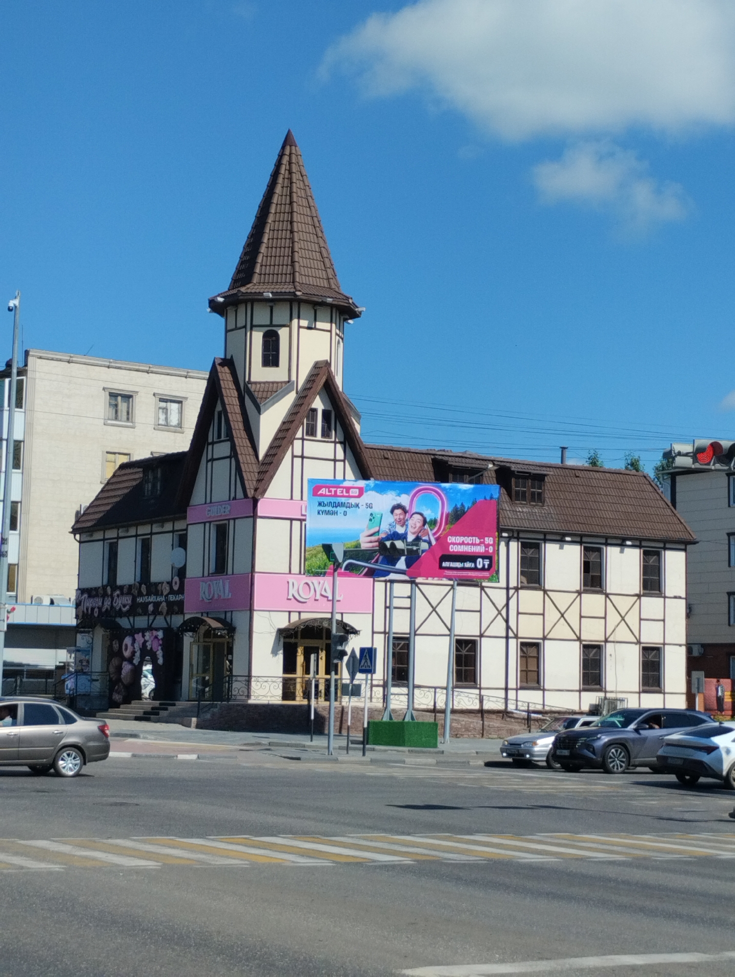 The image depicts a street scene in the city of Kostanay, Kazakhstan, on a sunny day with a clear blue sky and scattered clouds. The focal point is a distinctive building with a turret, constructed in a half-timbered architectural style, painted cream with brown trim. The building features multiple stories, a prominent brown tiled roof, and several windows. A large billboard advertising "ALTEL" mobile services is affixed to the side of the building, displaying images of two young men and promotional text. Below the billboard, pink signage with the word "ROYAL" is visible. Another sign above a doorway on the left side of the building appears to be in Cyrillic script and displays images of donuts. Several cars are present on the street, including a silver sedan driving away from the camera on the left and several other vehicles stopped at a crosswalk. The foreground shows a yellow and white striped pedestrian crosswalk. Traffic lights are visible in the upper right portion of the image. The general environment suggests a commercial or mixed-use urban area.