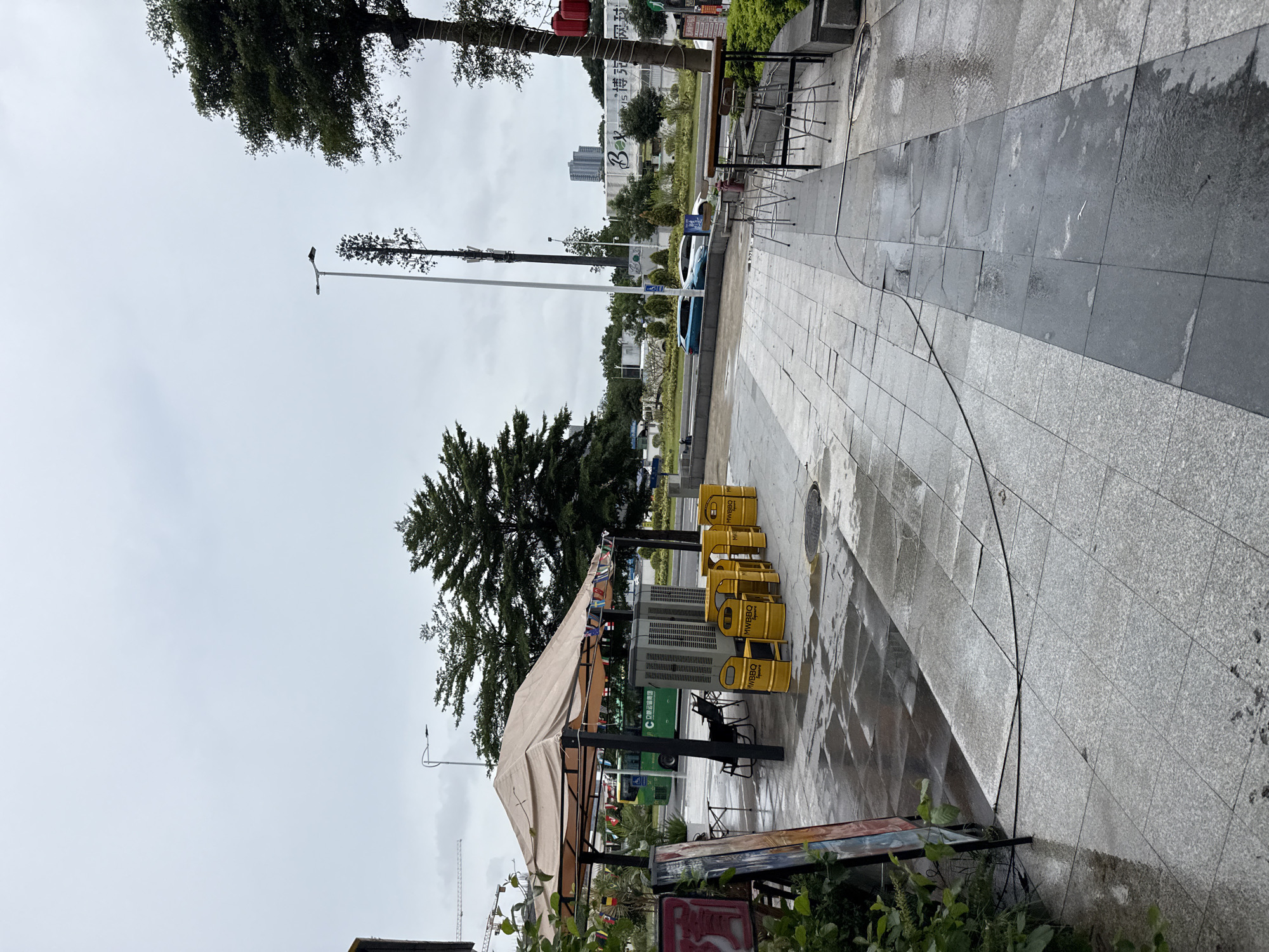 This is a sideways, eye-level outdoor shot of a sidewalk area with a covered seating area and several yellow trash bins. The weather appears to be overcast and the ground is wet, suggesting it might have recently rained or is currently drizzling.
The primary subjects are the yellow trash bins, which appear to be branded with "MWBBQ Square". There is a tent-like structure providing shade over a seating area with some chairs and tables. Behind the seating area, there's a green bus, and further in the background, there are buildings and trees. The sidewalk is paved with large, grey tiles, some of which are wet and reflective.
There's visible text in the image: "MWBBQ Square" on the bins, and "C立运输集团" and "B博克网球" on signs in the background. The overall scene suggests an urban environment, possibly a commercial area or a street with outdoor dining. The lack of people and the overcast sky contribute to a somewhat subdued or quiet atmosphere. The presence of trees indicates it's not a completely concrete jungle. The time of day is difficult to ascertain precisely due to the lighting conditions, but it appears to be daytime.