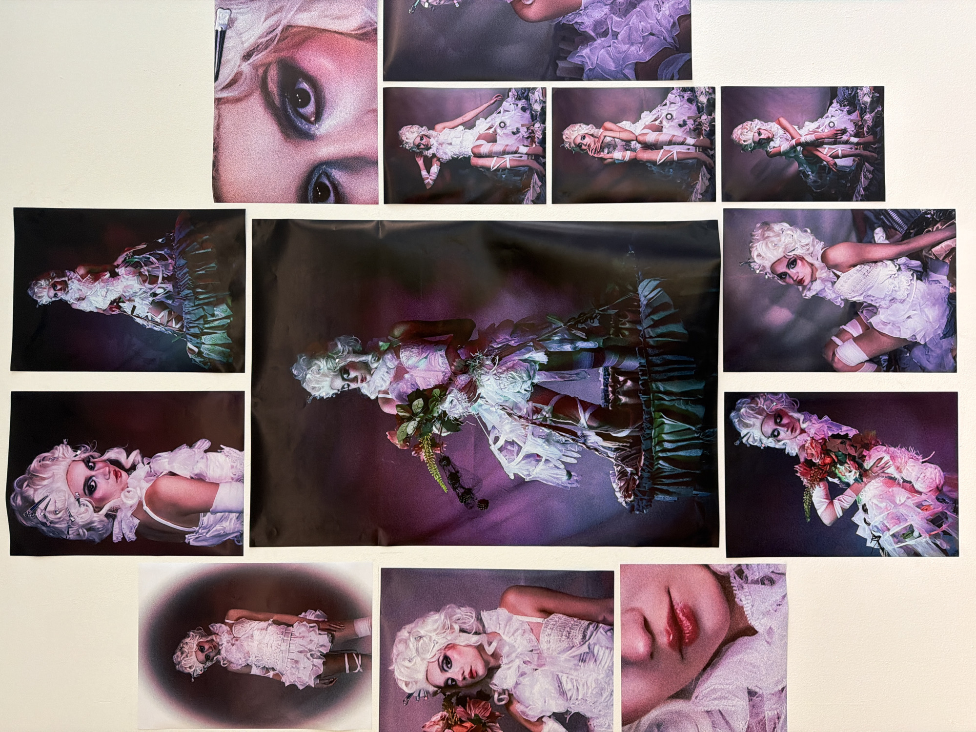 Multiple photographs are arranged on a light beige wall. The subject of the photographs appears to be the same individual, a person with pale skin, blonde curly hair styled in a wig, and heavy makeup, including dark eyeliner and pale lipstick. The person is wearing an elaborate white dress or costume with ruffles and frills. Some photos include props such as flowers and ribbons. The backgrounds vary but include shades of dark pink, purple, green, and black.