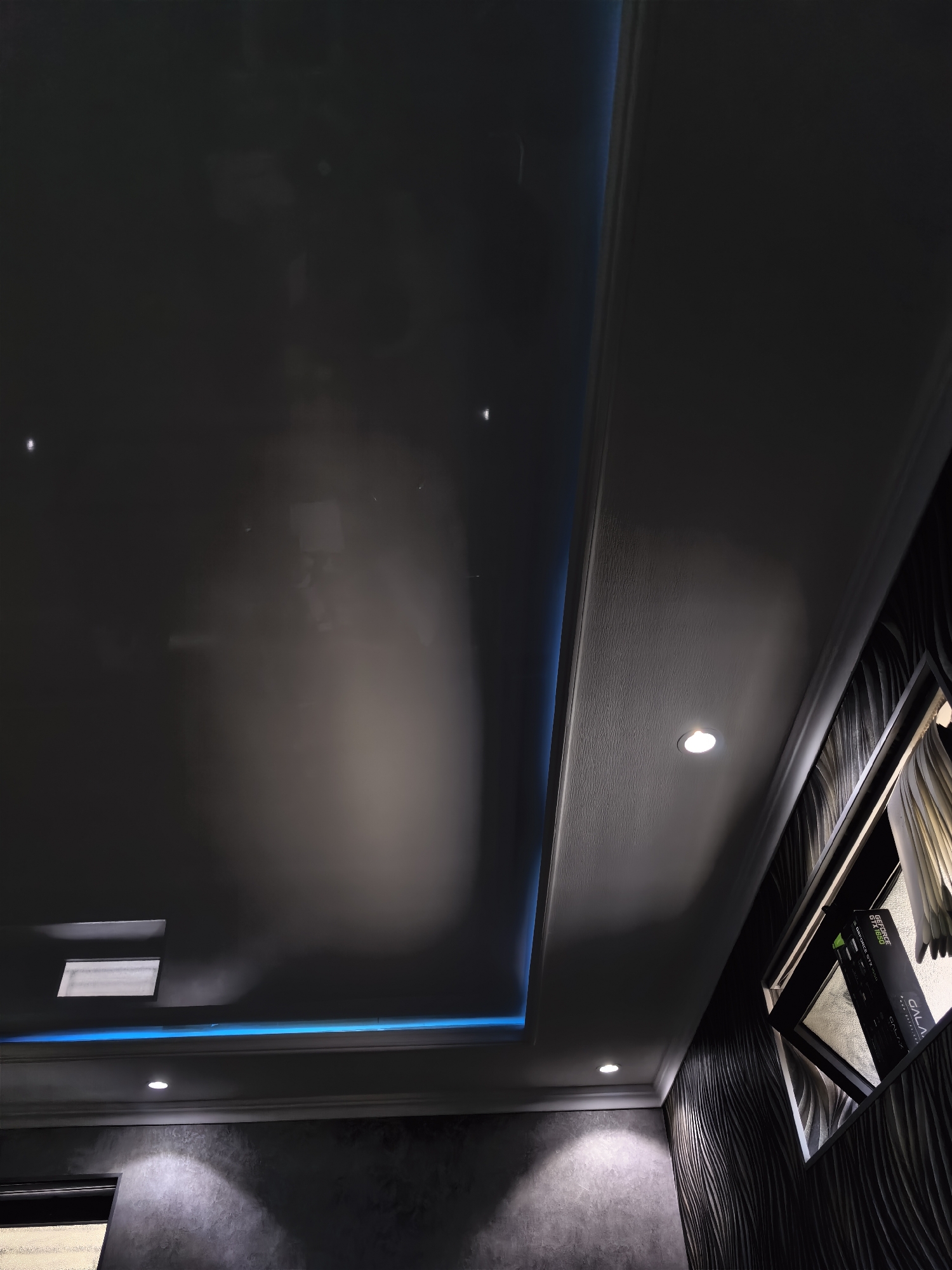 The image shows a modern interior ceiling design in Fergana City, Uzbekistan. The ceiling features a glossy black stretch ceiling with recessed lighting and blue LED strip lights. A portion of the ceiling is a textured white surface with a single circular spotlight. To the right, a window is visible, with what appears to be a computer graphics card box placed on the windowsill. The walls are decorated with dark, wavy patterned wallpaper. The lighting in the room is dim, with reflections visible on the glossy ceiling.