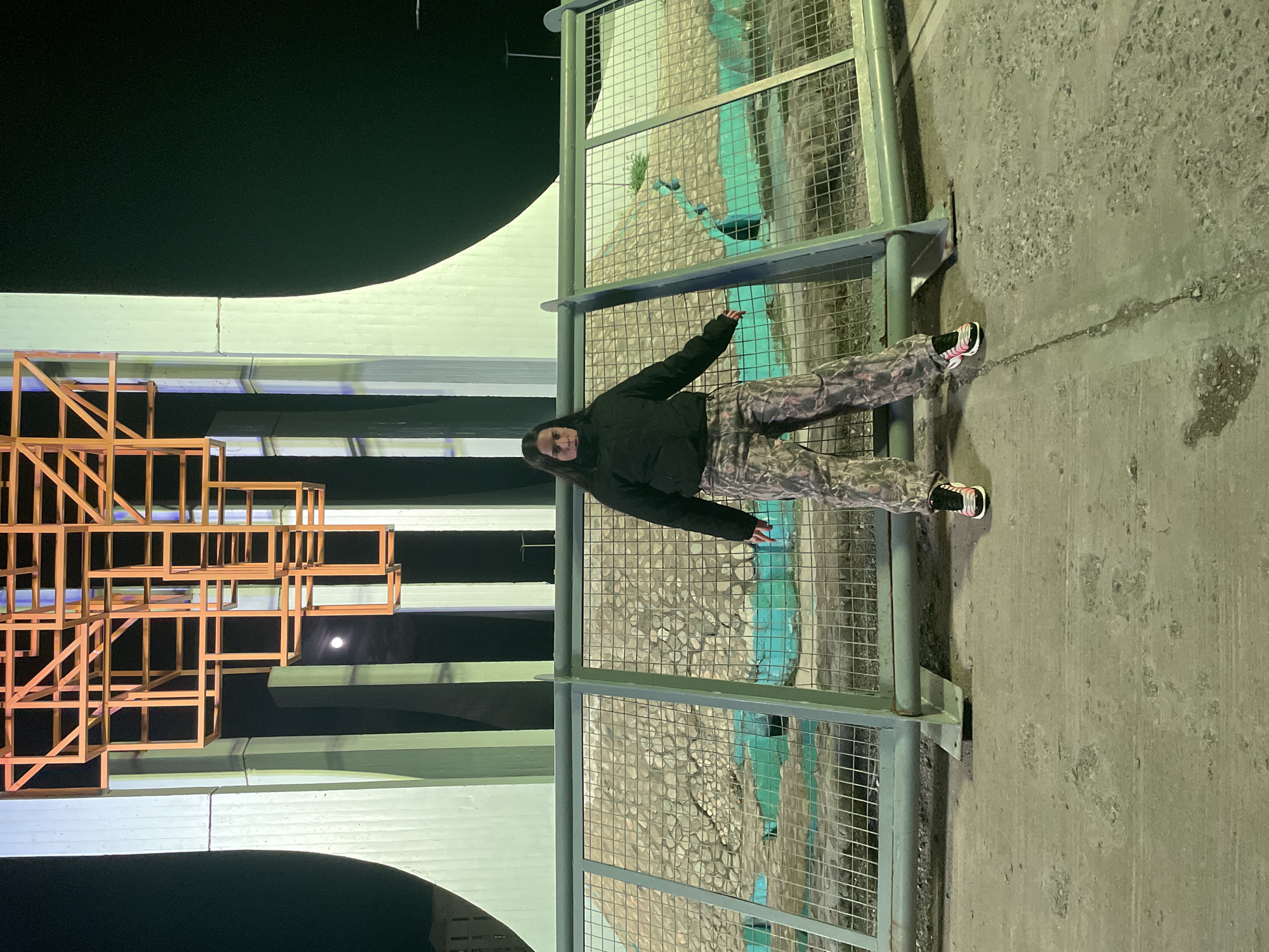 A woman stands between metal guardrails alongside a bridge, likely at night. She wears a black hooded jacket and patterned pants. The bridge structure includes a large, illuminated orange geometric sculpture. The environment appears to be urban. While the request mentions Ushuaia, Argentina, there's nothing in the scene to definitively confirm this location based solely on the visual information provided.