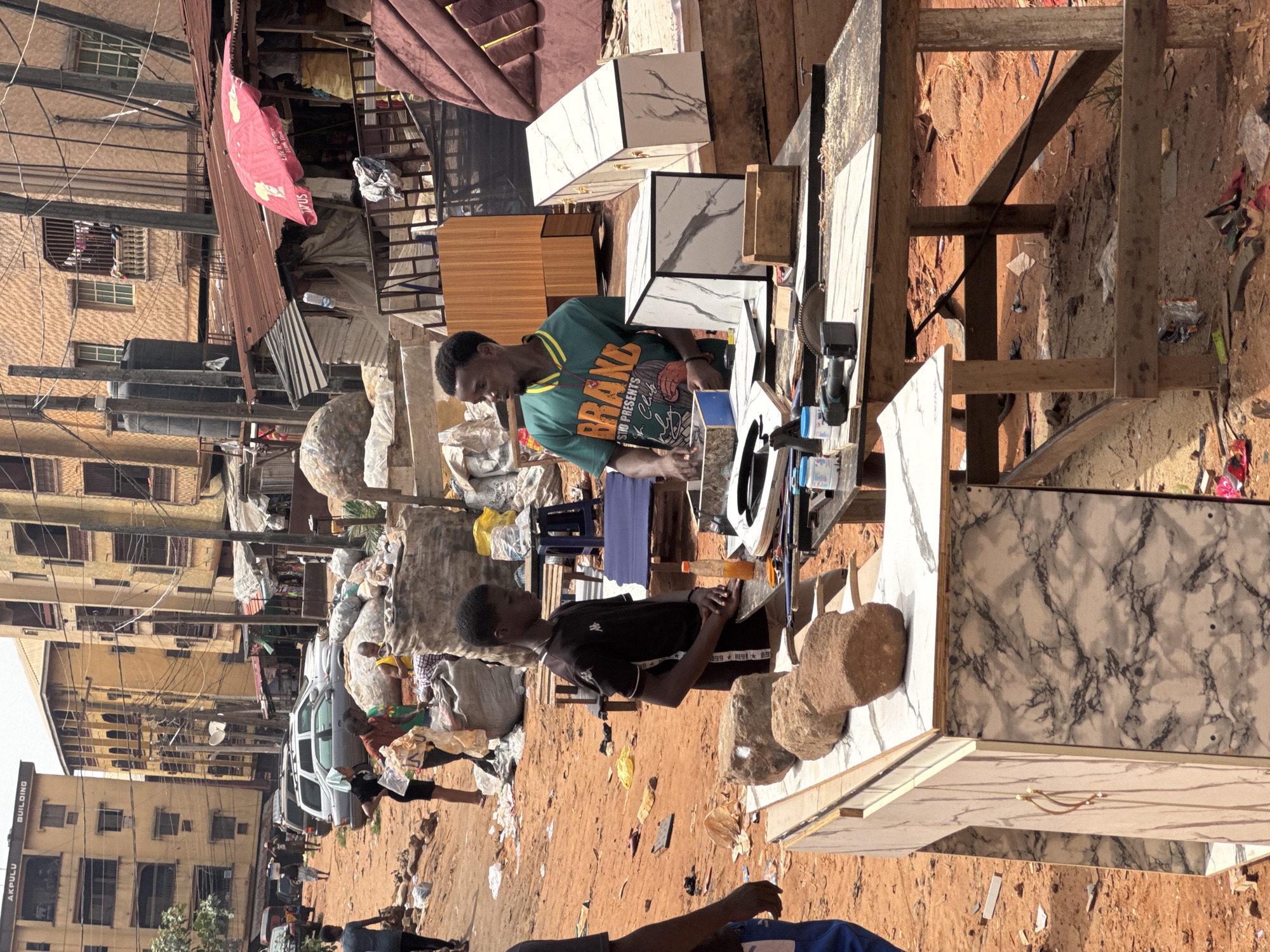 The image captures a street scene in Onitsha, Nigeria. Two men are centrally positioned, working on constructing or repairing furniture. One man wears a green t-shirt with the word "BRAND" prominently displayed and is operating a power tool, potentially cutting or shaping materials. The other man, in a black t-shirt, sits and appears to be observing or assisting. They are working on a makeshift wooden table covered with marble or marble-patterned tiles. Several finished or partially finished small cabinets or storage units are visible.
The environment is a bustling urban setting with buildings in the background, including one labeled "AKPULU BUILDING." Overhead power lines crisscross the sky. There are bags filled with materials, possibly recyclables or refuse, on the ground. A light-colored van is parked nearby. Several other people are walking around in the background, suggesting a busy market or commercial area. The ground appears to be primarily dirt or clay, common in the region. The overall impression is one of an informal economy and roadside business activity.