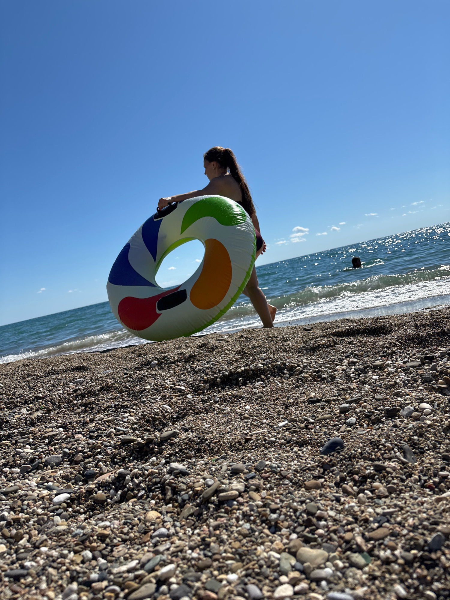A young female is walking on a pebble beach, holding a colorful inflatable ring. She is looking out at the sea, which is calm with small waves. The sky is clear and blue. In the background, a person is swimming in the water. The overall scene is peaceful and summery.