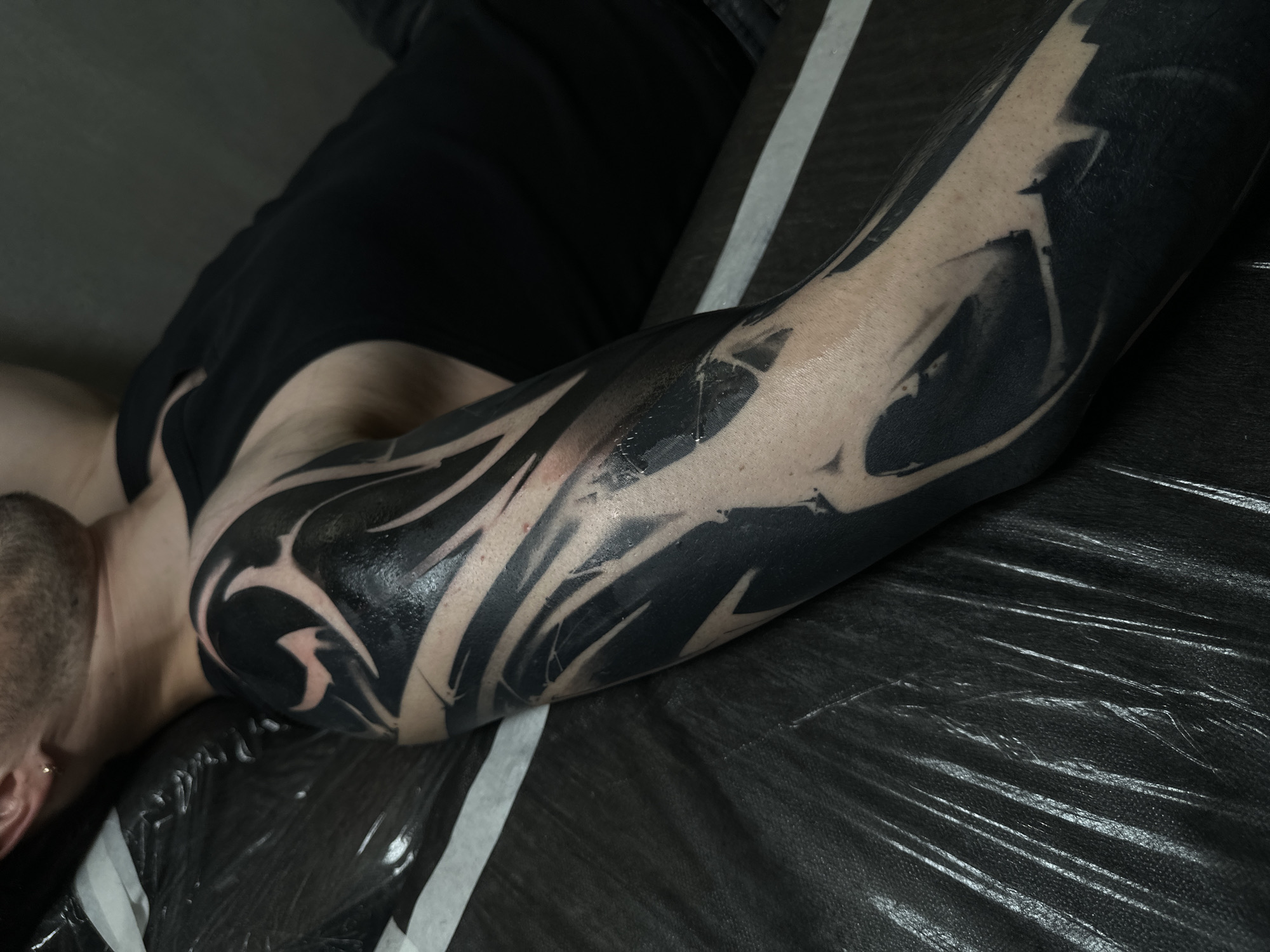 The image features a close-up of a person's left arm, heavily adorned with a blackout tattoo, in what appears to be an indoor setting, likely a tattoo studio in Moscow, Russia. The tattoo spans a significant portion of the arm, from the shoulder to the forearm, characterized by dense black ink that contrasts sharply with the person's fair skin. Within the blackout, negative space is skillfully utilized to create intricate, abstract designs, ranging from organic, flowing curves to more angular and sharp patterns. The surface of the tattoo appears fresh, exhibiting a glossy sheen, possibly from ointment or plasma after application.
The person is lying down, with only their left arm prominently displayed. A partial view of their head, including a shaved side, an ear with a small earring, and part of their neck and upper torso in a black t-shirt, is visible. The arm rests on a dark, textured surface, which seems to be a disposable plastic or vinyl covering, possibly over a tattoo chair or bed, and features distinct white stripes. The lighting is subdued, highlighting the intricate details of the fresh tattoo and its glossy appearance. There is no visible text present in the image.