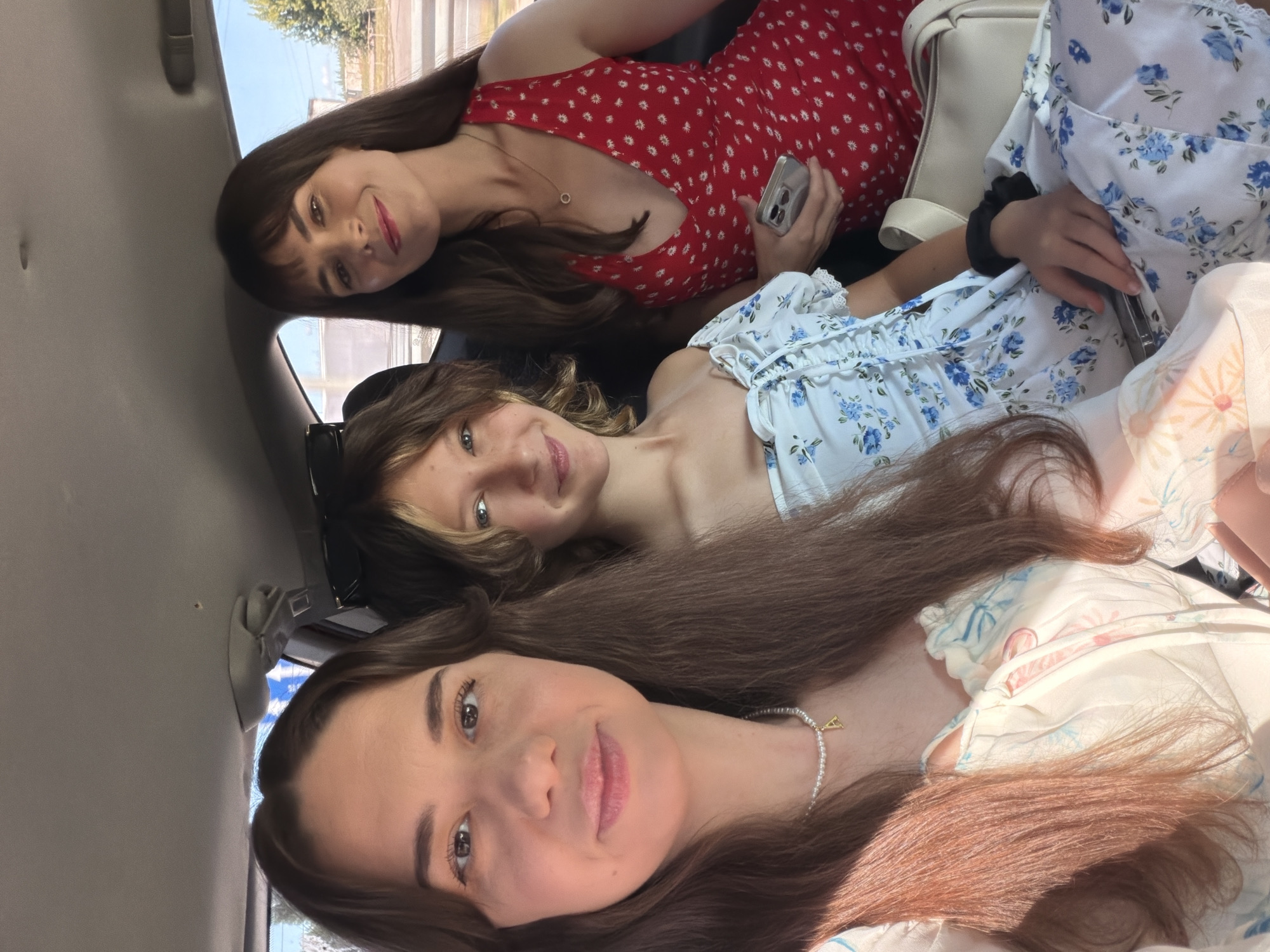 Three individuals are seated in the back of a car. The person in the foreground on the left is a woman with long brown hair, wearing a white dress with floral print and a pearl necklace. She is looking towards the camera. In the middle is a younger individual with light brown hair, wearing a blue floral dress. To the right is a woman with dark brown hair, wearing a red dress with white polka dots, and she is holding a phone. The scene appears to be daytime, with sunlight filtering into the car. The environment outside the car suggests a rural or suburban setting, possibly in Mali Kosharyshcha, Ukraine, given the presence of trees and a clear sky.