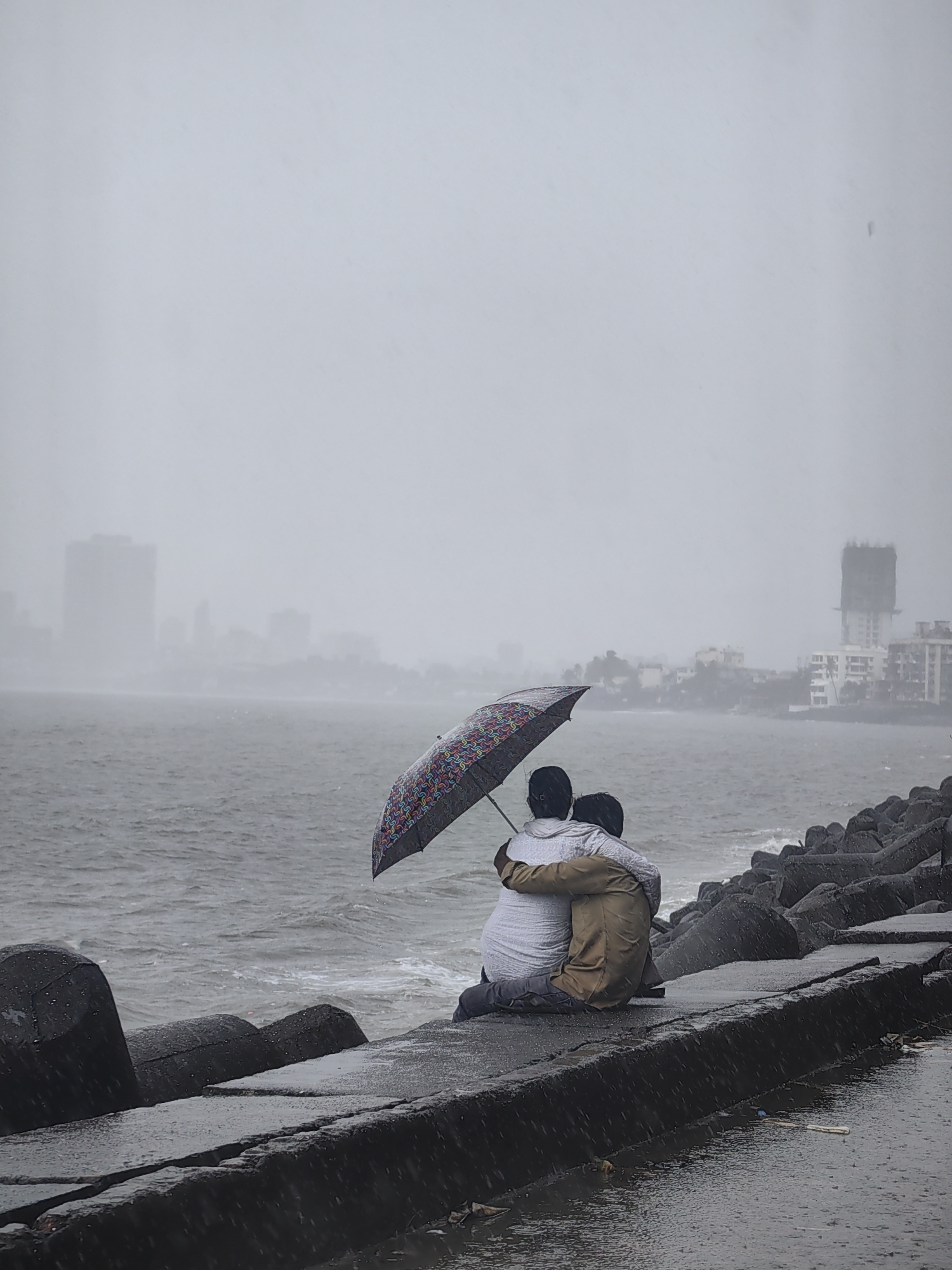 The image captures a couple embracing under a colorful umbrella on a rain-swept promenade, likely Marine Drive in Mumbai, India. The scene is overcast, with a hazy cityscape visible in the distance, featuring tall buildings shrouded in the mist of the monsoon. The choppy sea rolls against the Tetrapod breakwaters lining the shore, with raindrops visible in the air and on the wet concrete path. The couple, seated with their backs to the camera, are huddled together, seeking shelter from the downpour. The man, wearing a brown shirt and pants, has his arm around the woman, who is in a white top and dark pants. The umbrella, a focal point of the image, displays a vibrant, geometric pattern. The overall mood is one of intimacy and resilience against the elements.