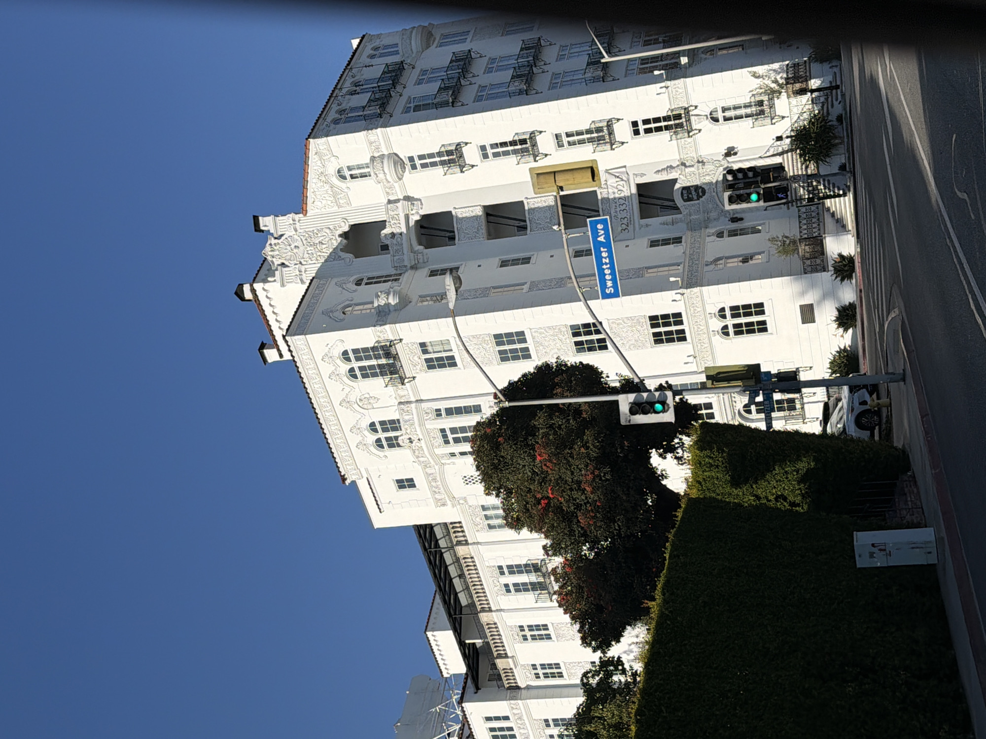 A wide shot shows a white, ornate building with a Mediterranean Revival architectural style under a clear blue sky. The building features detailed white ornamentation, arched windows, and balconies with wrought-iron railings. A lush green hedge lines the foreground on the left side of the image, and a street with a crosswalk runs in front of the building. A traffic light with a green signal is visible to the right of the building. A street sign reads "Sweetzer Ave" and a phone number is also displayed on a blue sign. A white car is parked on the right side of the street. A tree with red blossoms is situated in front of the building.