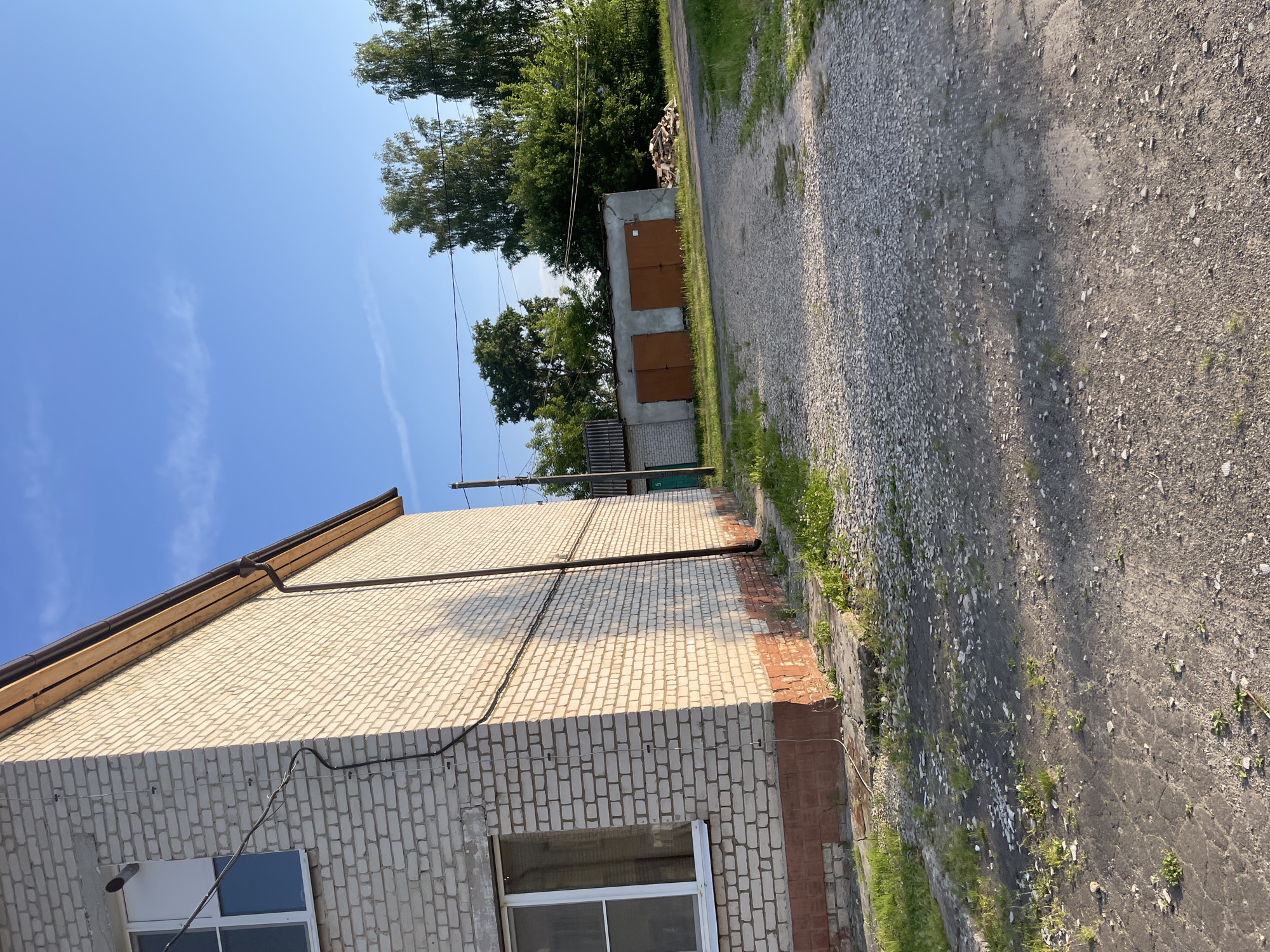 The image depicts an outdoor scene in the village of Prudý, Russia, under a clear blue sky with a few wispy clouds. In the foreground, a gravel and dirt pathway leads towards a small cluster of buildings. To the left, a section of a light-colored brick building with a red tiled base is visible, featuring a white-framed window. Wires are attached to the exterior of this building. Further down the path, a row of two garage doors, painted a muted orange, are situated in front of a low structure with a corrugated roof. To the right of the garages, a pile of chopped firewood is stacked. Beyond these structures, a line of green trees and foliage forms the background, with power lines running horizontally across the middle ground. The overall impression is of a rural or semi-rural residential area.
