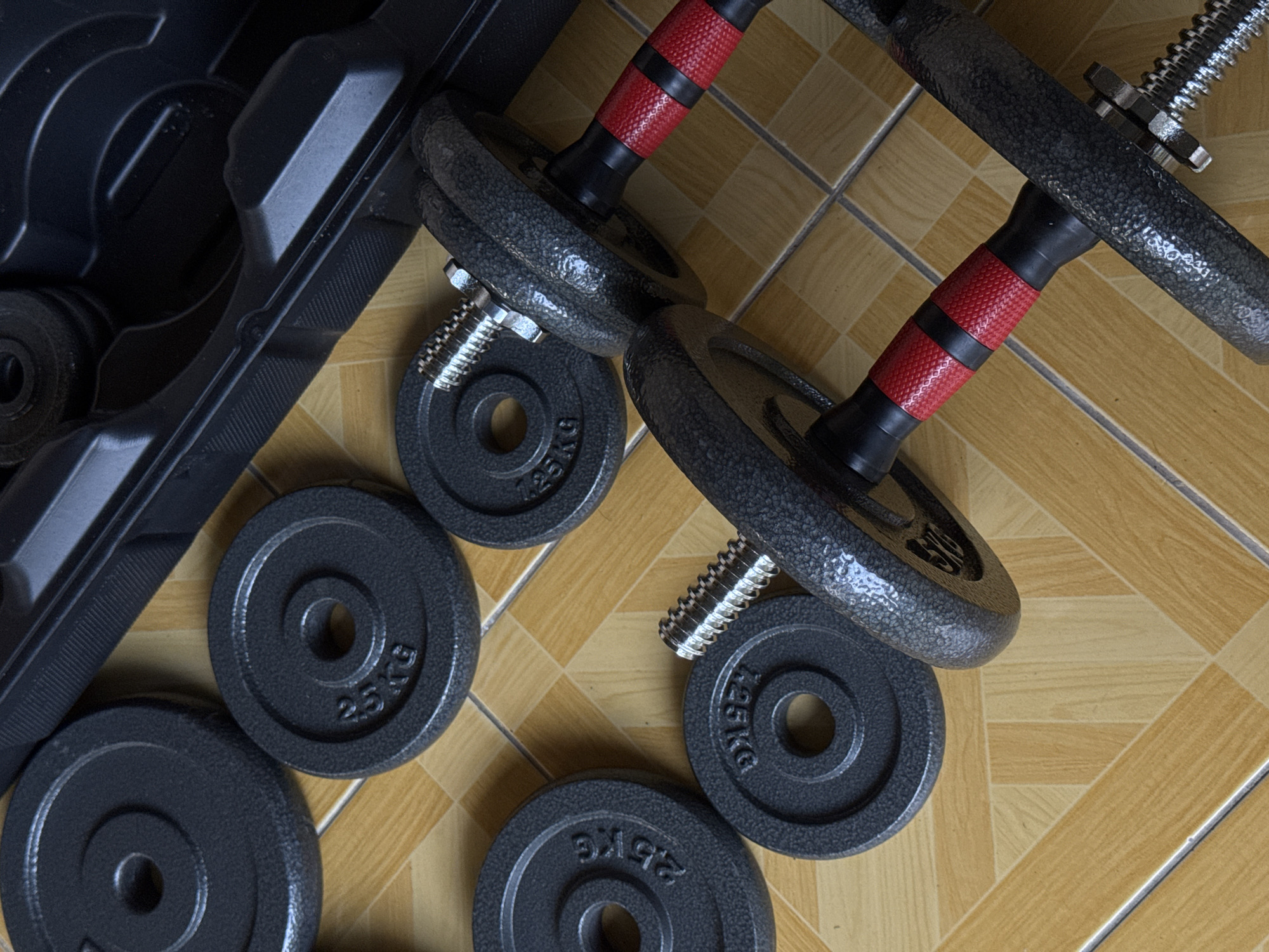 In Kalirancang, Indonesia, an overhead shot shows a set of dumbbells and weight plates arranged on a tiled floor. The dumbbells have black textured grips with red accents. Several round, black weight plates are scattered around the dumbbells, with some bearing the inscription "2.5 KG". A black plastic carrying case for the dumbbells is partially visible in the upper right corner of the image. The tiled floor has a pattern of light brown and beige squares.