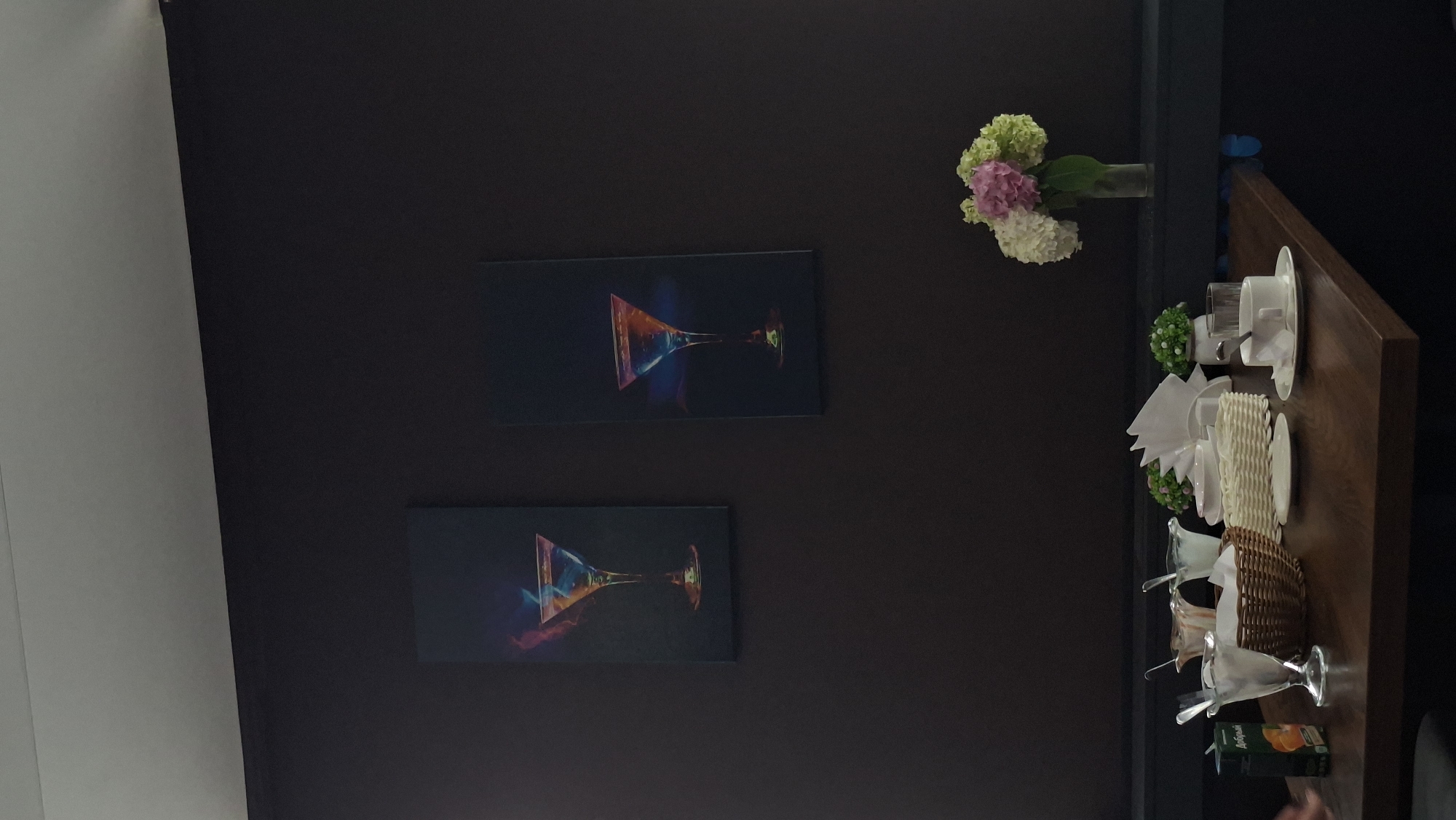 This image depicts a table setting in a dimly lit establishment, possibly a cafe or restaurant, in Myadzyel, Belarus. The dark walls provide a stark contrast to the objects on the table. Two framed art pieces featuring abstract representations of cocktails hang on the wall. In the foreground, a wooden table is laid out with several items. These include a carton of juice, three dessert glasses with spoons, a wicker basket, folded napkins, and a small vase with a bouquet of hydrangeas in shades of pink, white, and green. A white teacup and saucer with a spoon are also present, along with a clear glass. Two small artificial plants are placed on the table. The scene appears to be devoid of people.