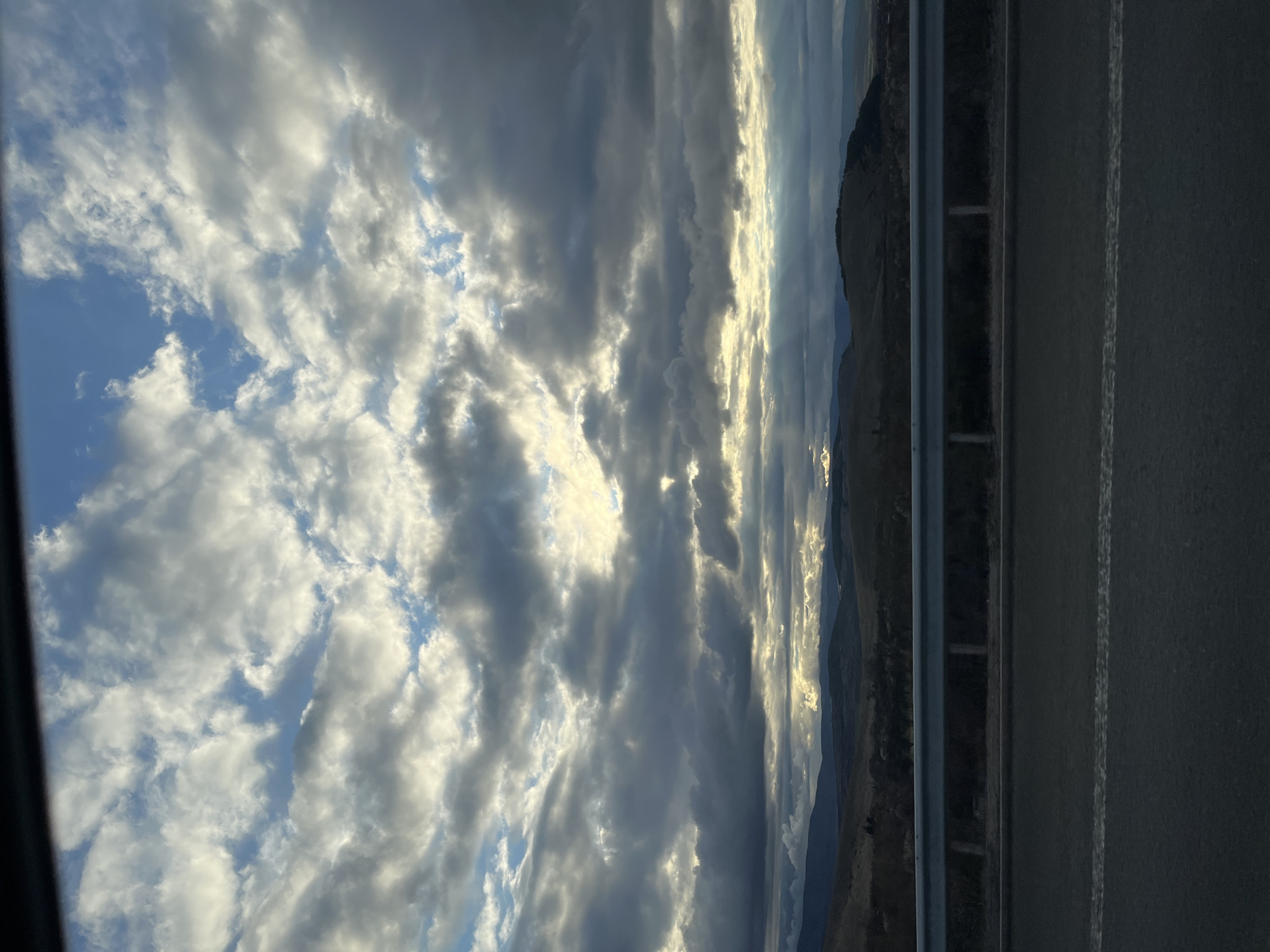 The image presents a view of a cloudy sky with patches of blue visible between the clouds. A mountain range is visible in the background, adjacent to a road with a guardrail. This is not identifiable with Staro Selo, Bulgaria.
