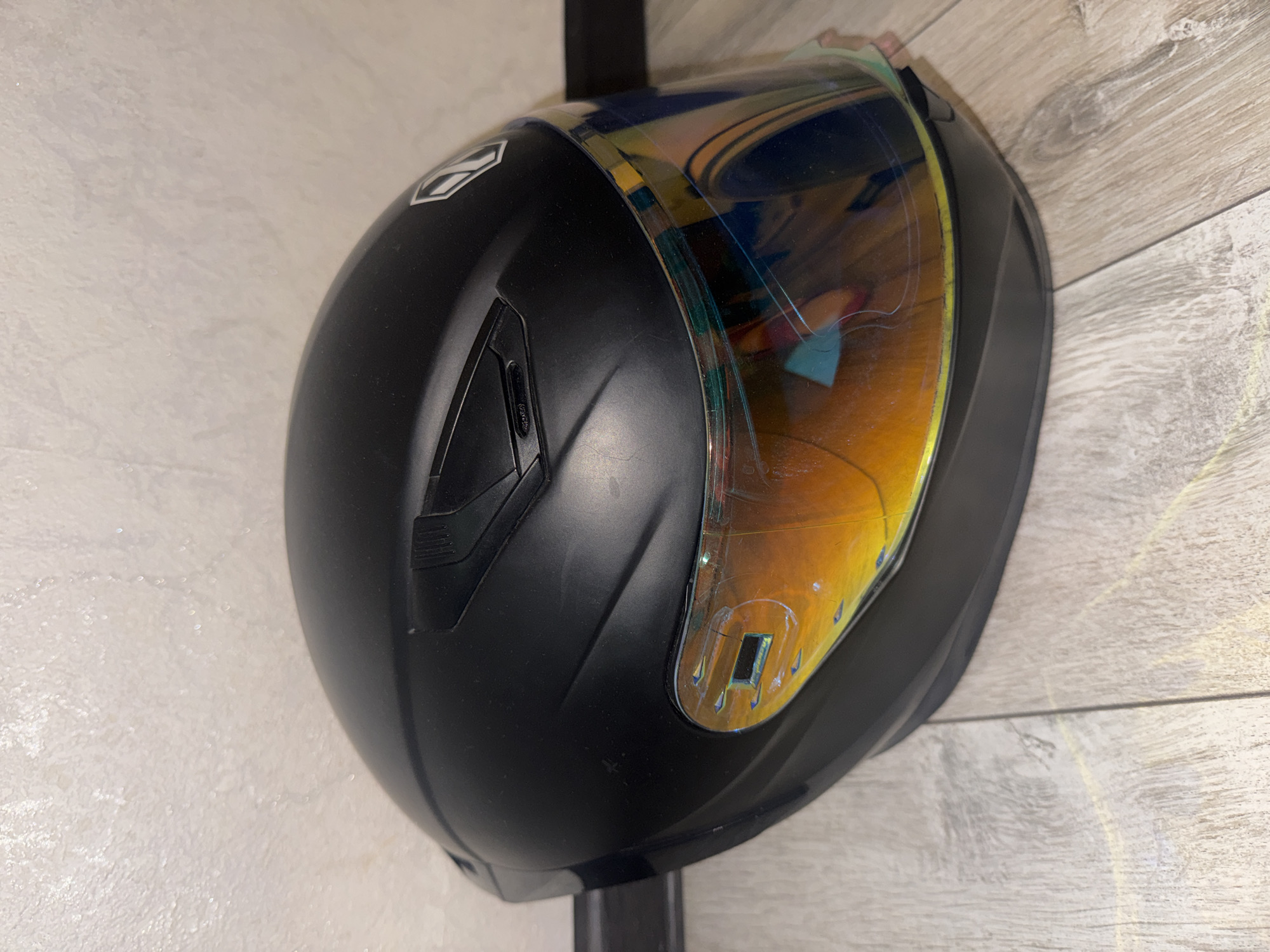 A black motorcycle helmet with an iridescent visor rests on a light-colored wooden floor. The helmet is positioned on its side, facing to the right. The background is a textured, light-colored wall. The scene is indoors, with no discernible urban or city elements visible, and therefore cannot be located in Bishkek City, Kyrgyzstan.