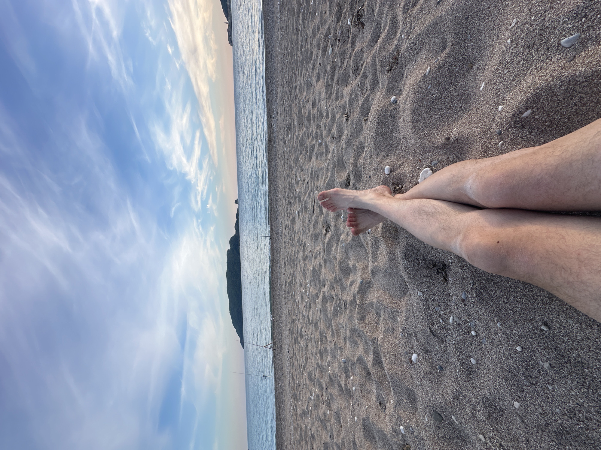A beach scene in Budva, Montenegro, features a sky with wispy clouds fading into the horizon above the water. The shore is made of dark, pebbly sand dotted with small white stones. In the foreground are two human legs stretched out on the sand, with bare feet crossed at the ankles. Two fishing poles are near the water's edge. A mountain rises on the horizon.