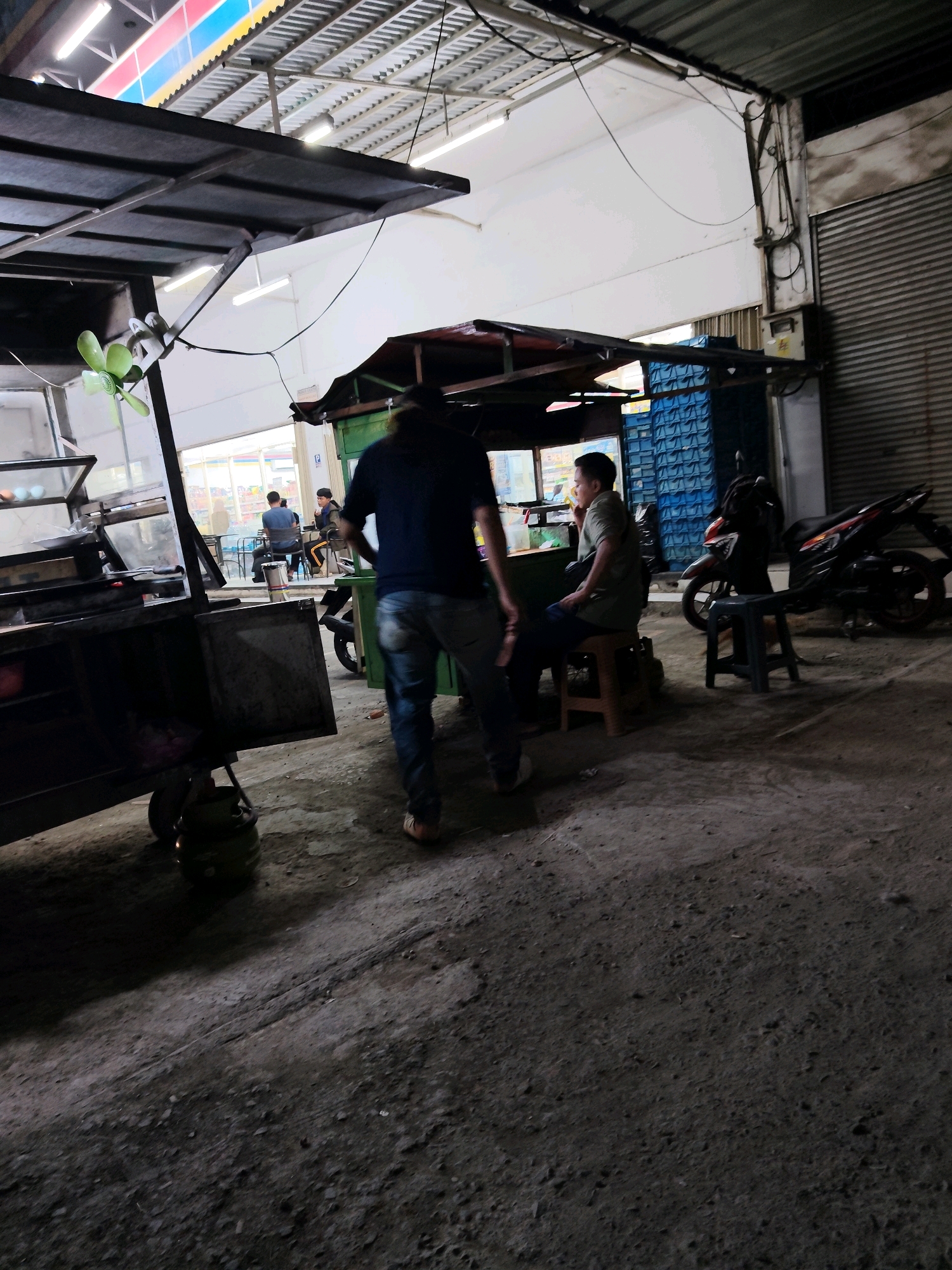 A medium shot captures an outdoor scene in Purwakarta, Indonesia, likely at dusk or night, illuminated by overhead artificial lights. In the foreground, a food cart with a dark metal frame and a green fan is partially visible. A person wearing a dark shirt and blue jeans is walking away from the cart, their back to the camera. To the right, a man in a green shirt and dark pants sits on a brown stool, facing a food stall with blue crates stacked behind it. A motorcycle is parked nearby. The ground is unpaved and appears to be dirt or gravel. The background shows a building with a corrugated metal roof and a closed shuttered door. The overall impression is of a casual, possibly commercial area.