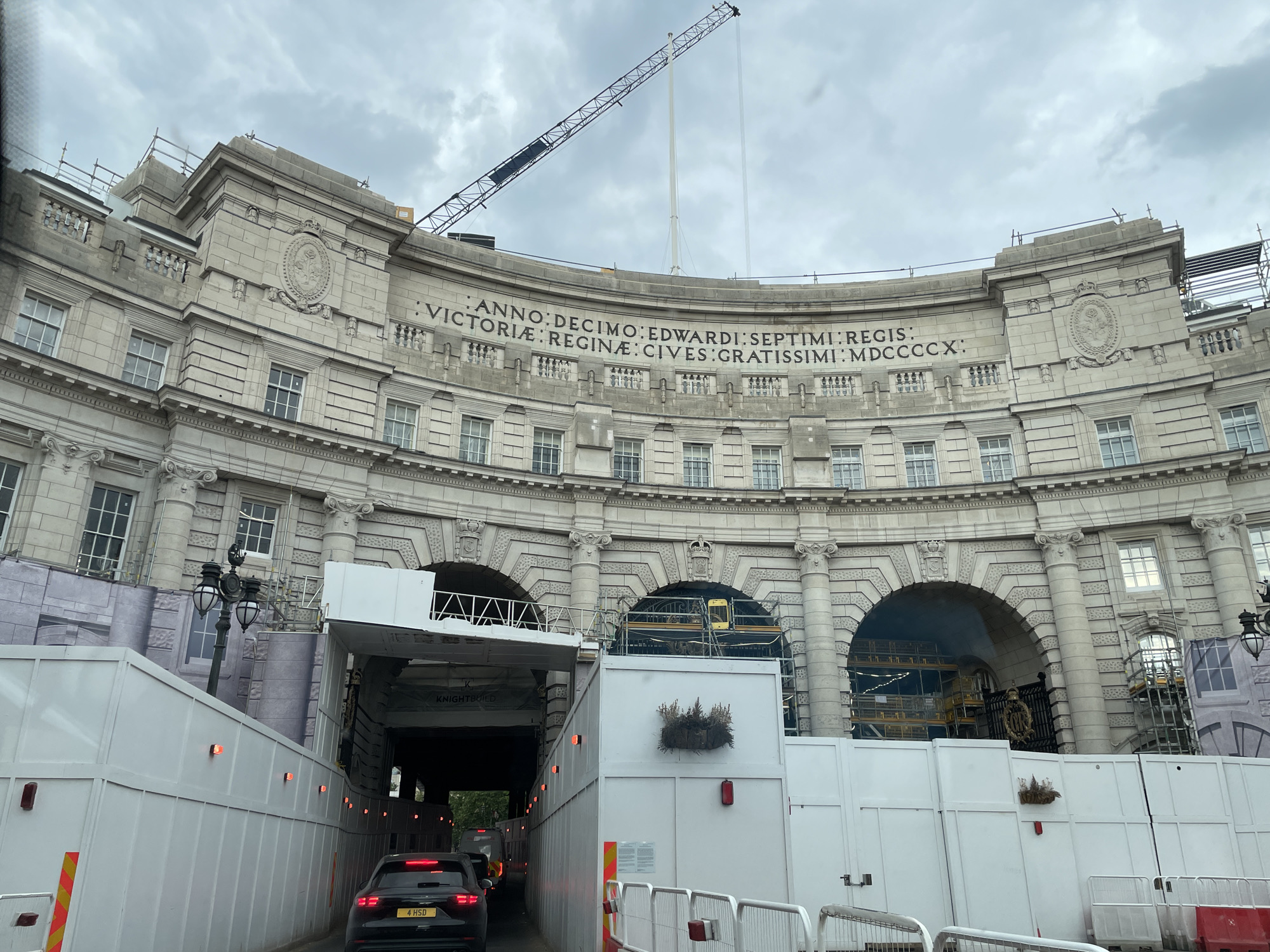 The image depicts a view from within a vehicle, looking towards Admiralty Arch in London, United Kingdom. The arch is partially obscured by white barriers, presumably for construction or maintenance work. A black car with license plate "4 HSD" is in the foreground, moving through a temporary tunnel created by the barriers. A crane is visible extending above the arch. The inscription "ANNO:DECIMO:EDWARDI:SEPTIMI:REGIS:VICTORIÆ:REGINÆ:CIVES:GRATISSIMI:MDCCCCX:" is inscribed on the arch.