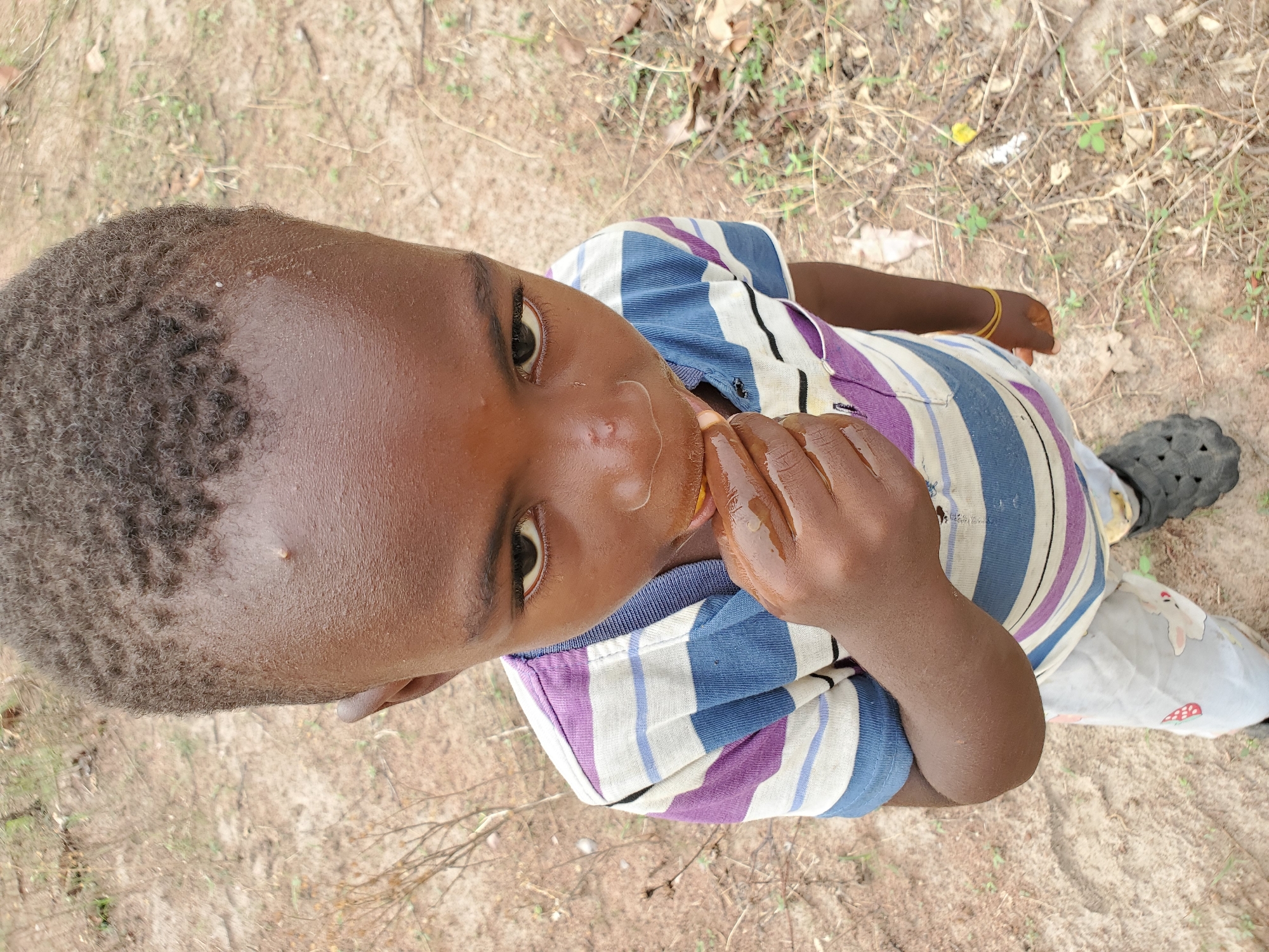 An overhead, eye-level shot captures a young child of African descent looking directly up at the camera in an outdoor setting in Mararaba, Nigeria. The child has short, curly dark hair and dark skin, and appears to be a boy. He is wearing a multi-colored striped short-sleeved shirt with blue, purple, white, and black stripes, and a yellow elastic bracelet on his right wrist. The child is actively eating a bright yellow-orange fruit, possibly a segment of orange or mango, which he holds in his hands. His face and hands appear somewhat wet or sticky from the fruit. The background consists of dry, light brown earth with scattered dead leaves, twigs, and sparse green weeds, suggesting a dry and perhaps rural environment. Part of a black, possibly Croc-style, shoe is visible on his left foot, and light-colored pants with a subtle pattern are also partially visible. The lighting indicates it is daytime. The child's expression is neutral and engaging as he meets the viewer's gaze. There is no visible text in the image.