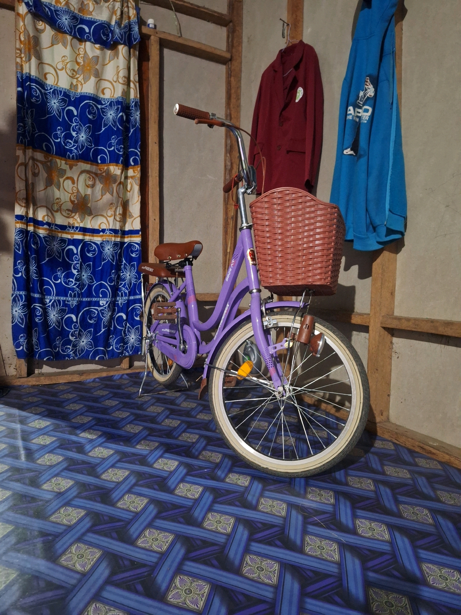 A lavender bicycle with a brown wicker basket is parked indoors on a blue patterned floor. The bicycle has a brown seat, brown handlebars, and a brown lamp attached to the front. To the left of the bicycle, a blue and white patterned curtain hangs. Behind the bicycle, a maroon jacket and a blue hoodie are hung on a wooden frame against a light-colored wall. The scene appears to be a storage area or a room within a residential building in Air Tawar, Indonesia. There are no people present in the image.