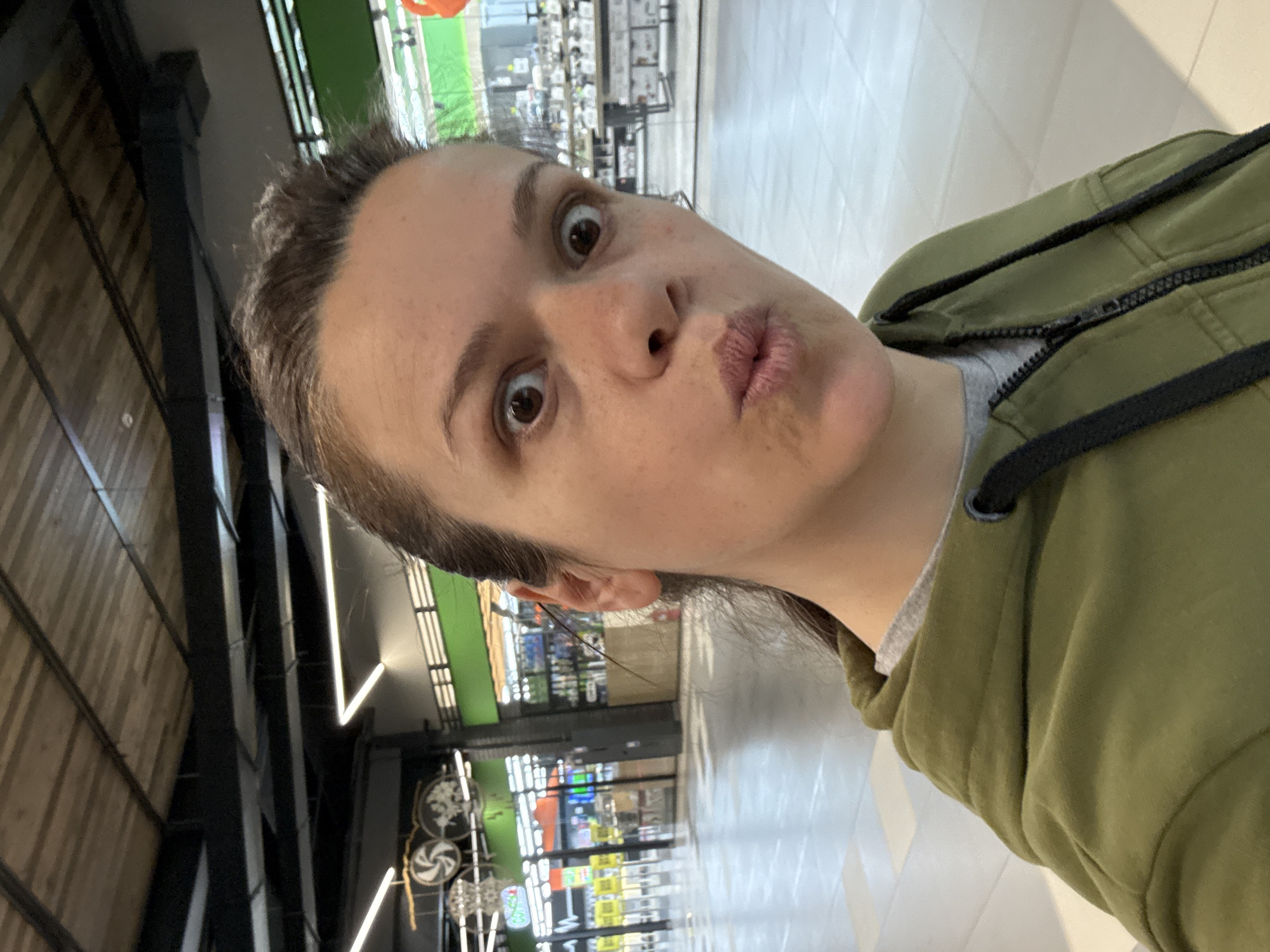 A woman is taking a selfie in a supermarket. She is wearing a green hoodie and making a surprised face with pursed lips. In the background, supermarket aisles are visible with shelves stocked with various products. A sign with decorations hangs from the ceiling. The image appears to be taken in Hatne, Ukraine based on the tags.