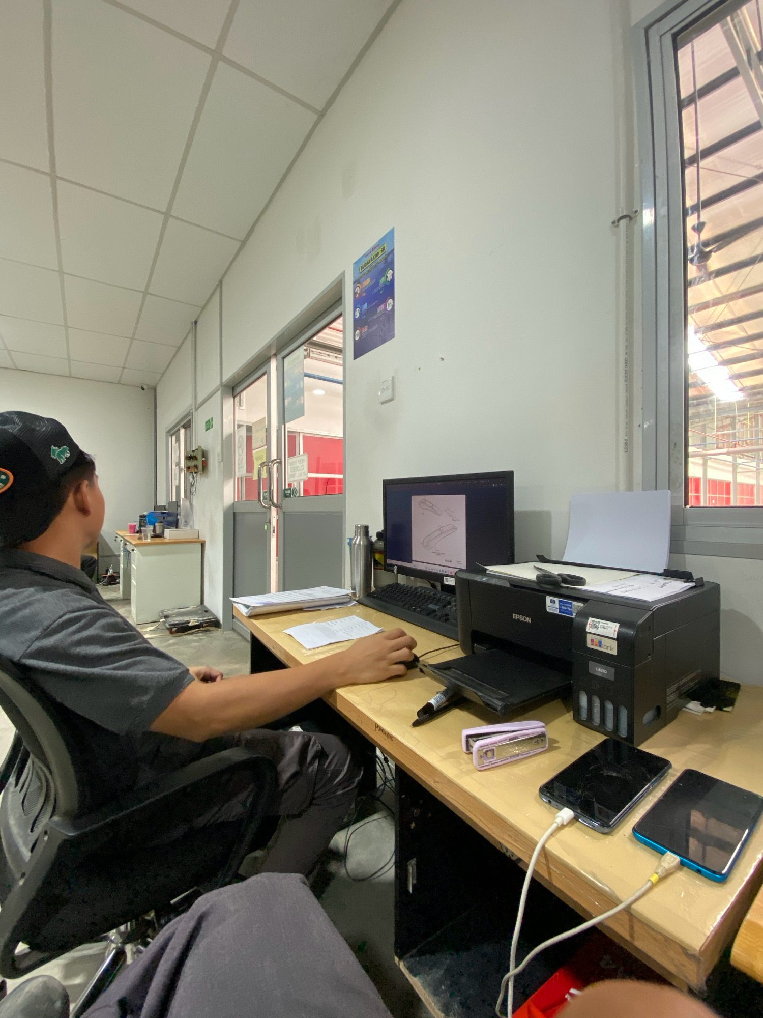 The image depicts an indoor scene, likely an office or workshop environment, located in Batam, Indonesia. A person wearing a black cap and a grey polo shirt is seated at a wooden desk, facing away from the camera and towards a computer monitor. The monitor displays a technical drawing. In front of the monitor is an Epson printer with paper loaded. Also on the desk are a stapler, two mobile phones charging, a water bottle, and some papers. To the left of the desk, there's another workstation with a filing cabinet and what appears to be a scale on the floor. The room has a tiled ceiling with fluorescent lighting and large windows revealing an industrial or construction setting outside. The overall impression is one of a functional workspace.