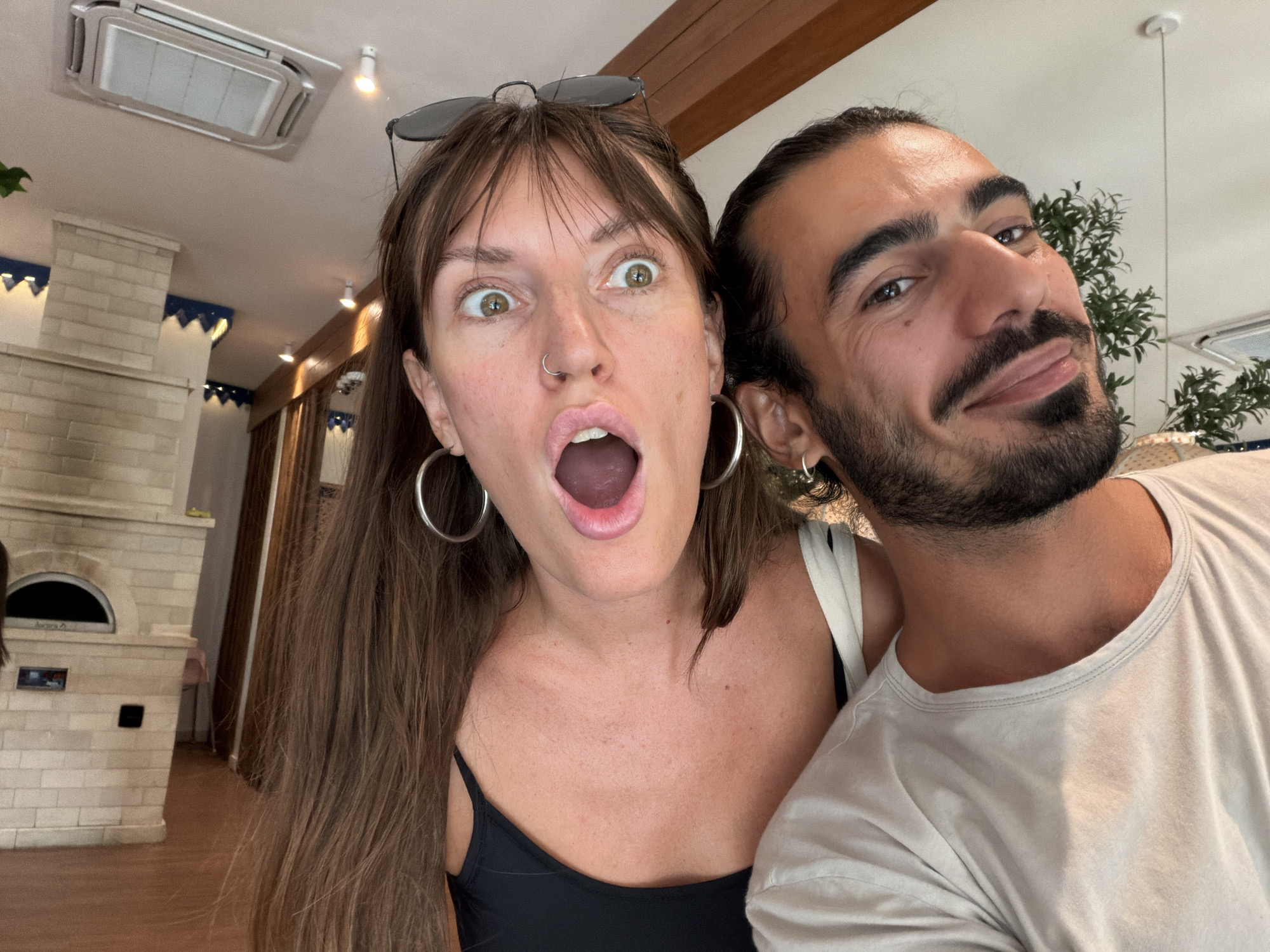 This media file is a selfie taken by two people, a woman and a man, in what appears to be a restaurant or cafe. The woman is in the foreground, with her mouth open in a surprised expression, wide eyes, and hoop earrings. She has long brown hair and is wearing a black top. The man is to her right, with a slight smile and a beard. He has dark hair pulled back and is wearing a light-colored t-shirt.
Behind them, the setting is a well-lit interior with wooden beams on the ceiling and a brick structure that resembles a pizza oven. There are also glimpses of plants and what looks like air conditioning units on the ceiling. The overall ambiance suggests a casual, possibly warm environment. The lighting indicates it is daytime. The emotional expressions on their faces suggest they are having fun or are surprised by something, possibly posing for a picture.
No visible text is present in the image. The location context is North Kuta, Indonesia.