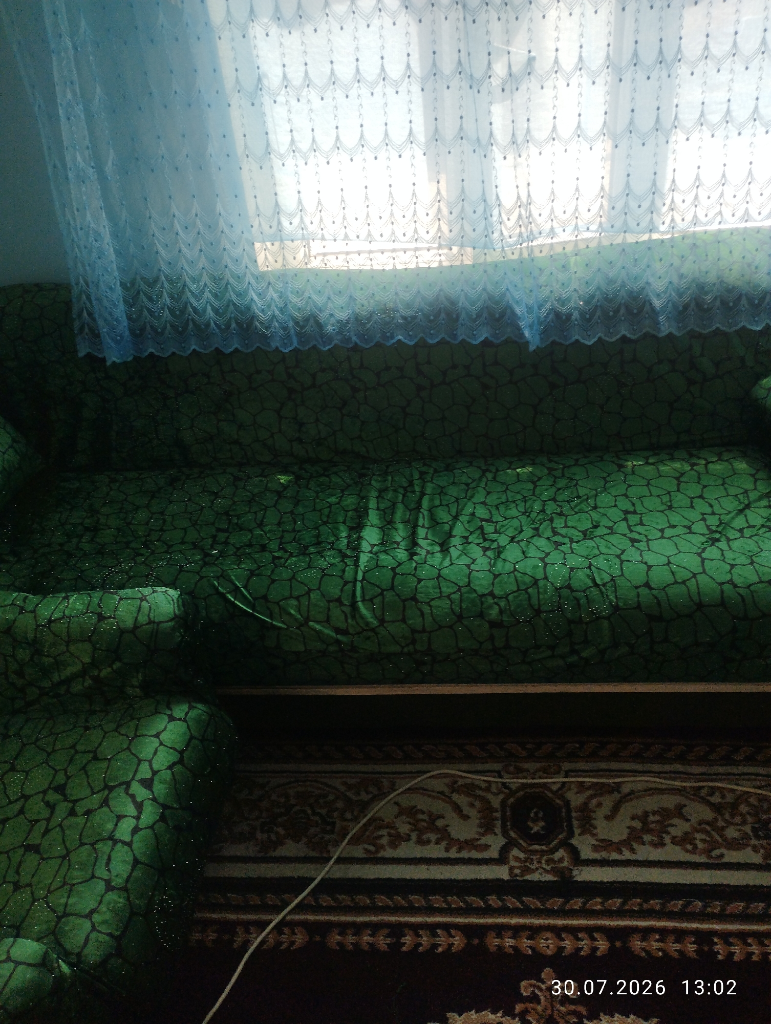 A green sofa with a patterned cover is in the foreground. A blue lace curtain hangs in front of a window behind the sofa. A carpet is on the floor in front of the sofa. A white cable is visible on the carpet. The date stamp "30.07.2026 13:02" is in the lower right corner of the image. The scene is indoors, likely a living room in a residence in the city of Quyosh, Uzbekistan.