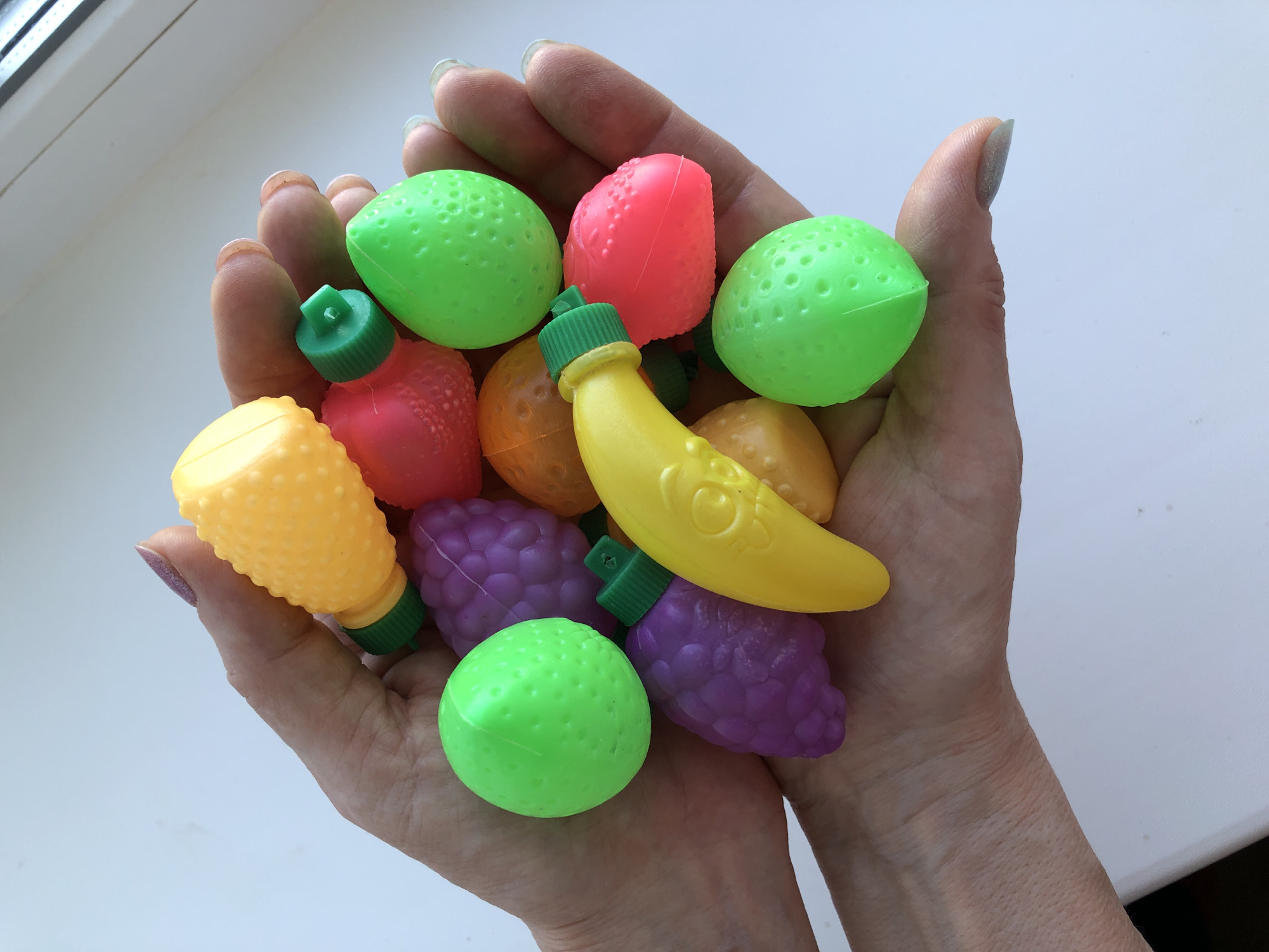 A pair of hands is holding several colorful plastic toy fruits. The fruits include: three green citrus fruits, two purple grape bunches, one red strawberry, one orange citrus fruit, one yellow pear, and one yellow banana. The fingernails of the hands are long and painted with a glittery silver-gray polish. The background is a plain white surface. The location is not discernable from the image.
