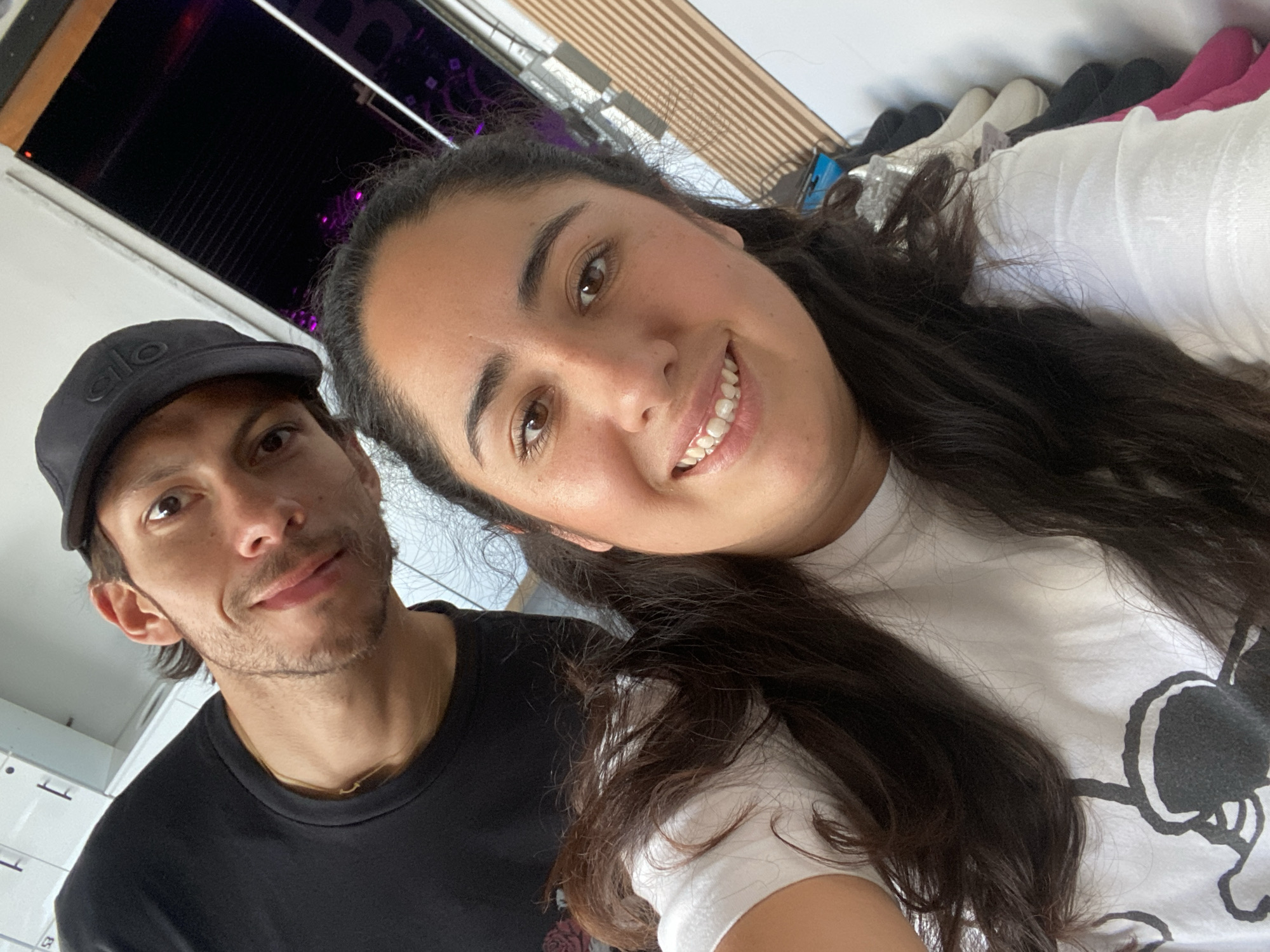 The image captures a selfie of two individuals indoors. A man with short dark hair, a slight mustache, and wearing a black baseball cap and black t-shirt is on the left. A woman with long, dark wavy hair, wearing a white t-shirt, is on the right. They are both smiling. Behind them, partially visible, is a window with what appears to be horizontal blinds or slats. In the background, there are also some items that look like shoes and clothing. There is no location information available from the image to confirm that it was taken in the city of Puebla, Mexico. The image provides no information related to actions or interactions that extend beyond the pose.