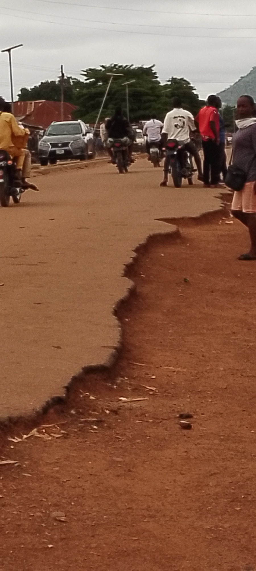 The image depicts a street scene in Gboko, Nigeria, with several individuals on motorcycles. The road surface is made of reddish-brown earth and shows signs of erosion along its right edge, creating a sharp drop-off. In the background, a dark-colored SUV is visible, along with a building with a red corrugated metal roof. Trees with dense green foliage line the background, and a power line stretches across the upper portion of the image. The sky is overcast.
On the road, a person in a yellow shirt is seated on a motorcycle on the left side of the frame. Further down the road, a person in a black shirt is riding a motorcycle, and another person in a white shirt is on a motorcycle behind them. To the right, a person in a red shirt is standing, and a woman in a grey shirt is walking away from the camera. The motorcycles appear to be common commuter models. The people's clothing suggests a warm climate. The overall impression is of a typical day in a busy urban or semi-urban area in Nigeria, with motorcycle traffic being a prominent mode of transportation.