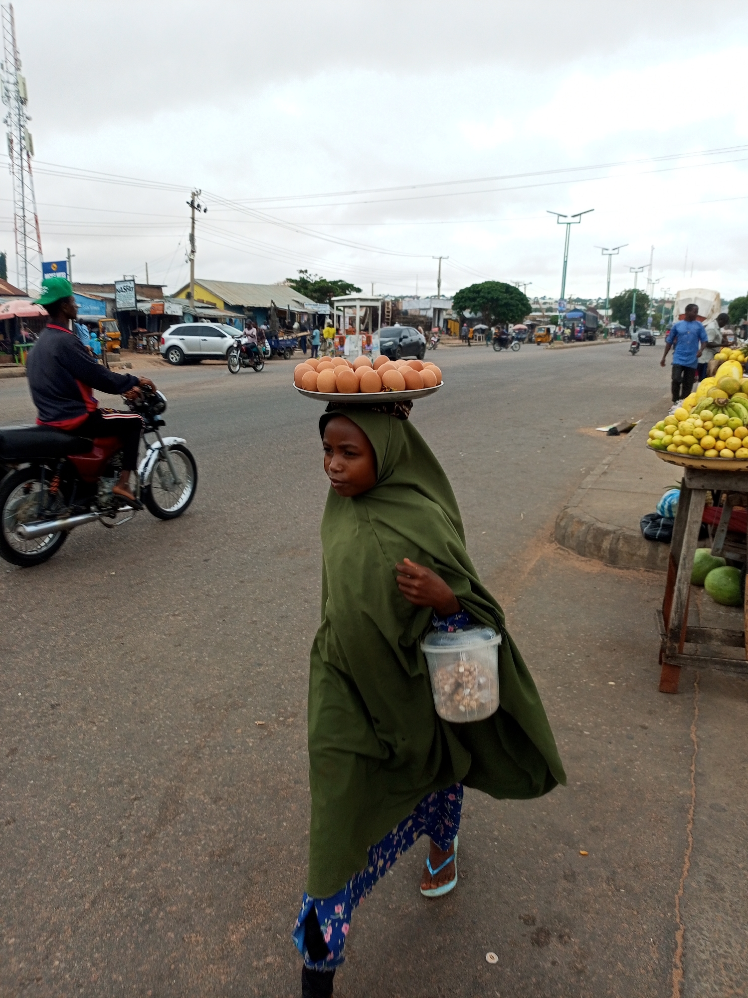 In Gombe, Nigeria, a young person wearing a green hijab and a dark green cloak walks down a paved street. They are carrying a large plate piled with brown eggs balanced on their head, and a clear plastic container filled with nuts in their right hand. They are wearing blue and white patterned leggings and light blue flip-flops. In the background, a person on a red motorcycle is seen from the side, wearing a blue jacket and a green cap. A silver car is parked behind them, and market stalls with fruit and other goods are visible on the right side of the road. The sky is overcast, and the scene appears to be a busy street in a market area.