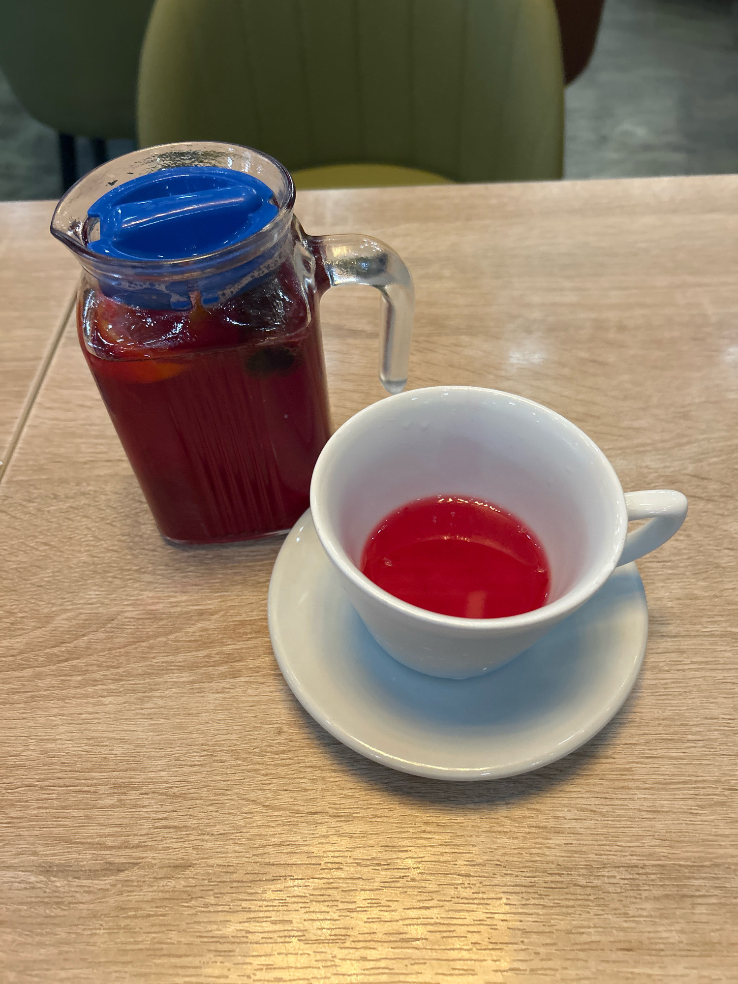 A glass pitcher filled with a crimson beverage and fruit slices, topped with a blue lid, sits on a wooden table next to a white teacup and saucer containing the same red liquid. The scene is indoors, likely in Moscow, Russia, with a blurred green chair visible in the background. The lighting is warm, suggesting an indoor setting.