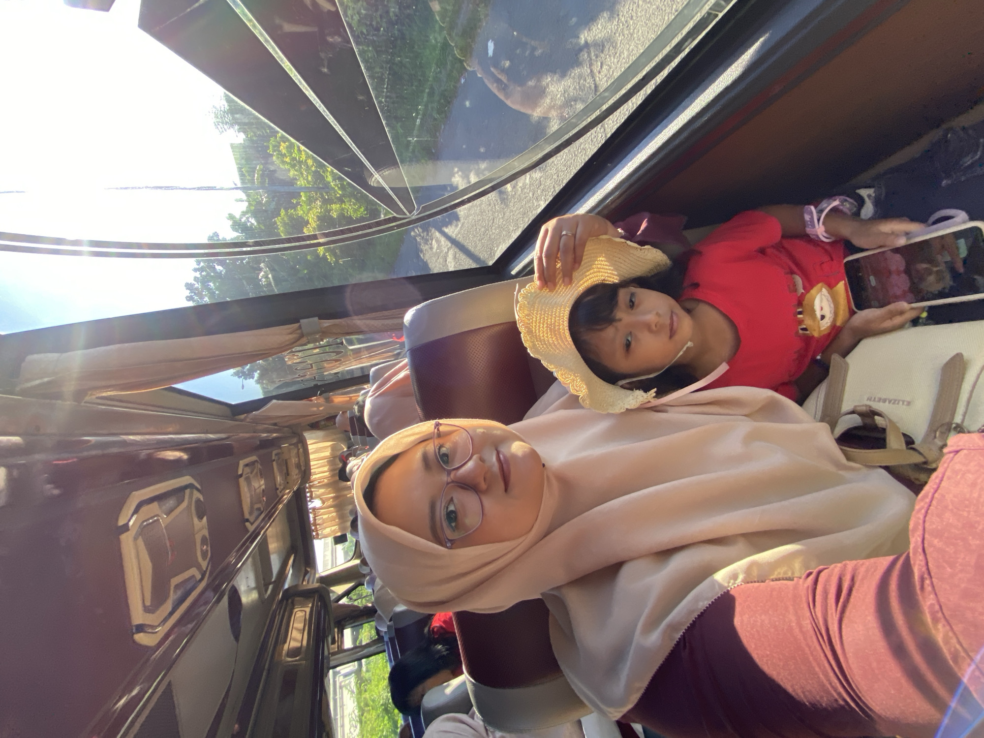 An adult female and a young girl are photographed inside a passenger bus in Kebonagung, Indonesia. The adult female, positioned on the left, wears a light beige hijab and maroon clothing, with purple-framed eyeglasses, and displays a slight smile. She appears to be taking the photograph with a smartphone. The young girl, seated to her right, wears a red t-shirt with a yellow cartoon character graphic and a light-colored straw hat secured by a chin strap. She holds a white smartphone with both hands, displaying an image on its screen, and has a neutral expression. Sunlight streams into the bus from the right, illuminating the interior and creating lens flare effects.
The bus interior features maroon and beige seats, and maroon overhead compartments with integrated lighting units. Light brown curtains are drawn along the windows. Through the windows, a paved road, green trees, and bushes are visible, indicating an outdoor environment during daylight hours. Distant structural elements of what appears to be an overpass or elevated road are also visible through the upper portions of the windows. A light-colored bag with "ELIZABETH" printed on it rests near the adult female's lap.