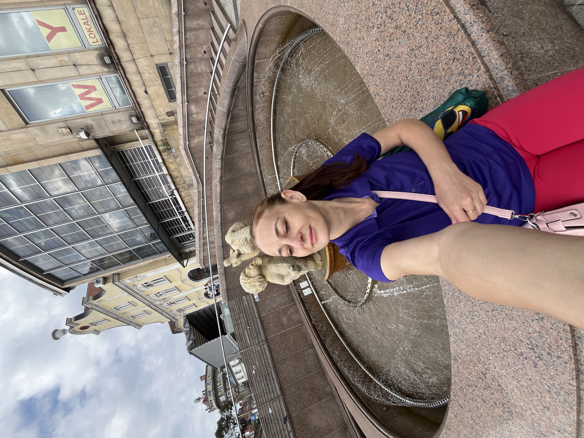 A woman in a purple shirt and pink pants takes a selfie in front of a fountain in an outdoor urban setting. The fountain features a small sculpture of cherubs. Behind the fountain, there are stone steps and railings leading up to buildings. One building is yellow with traditional architecture, and another is a modern building with large windows and signage. The sky is cloudy.